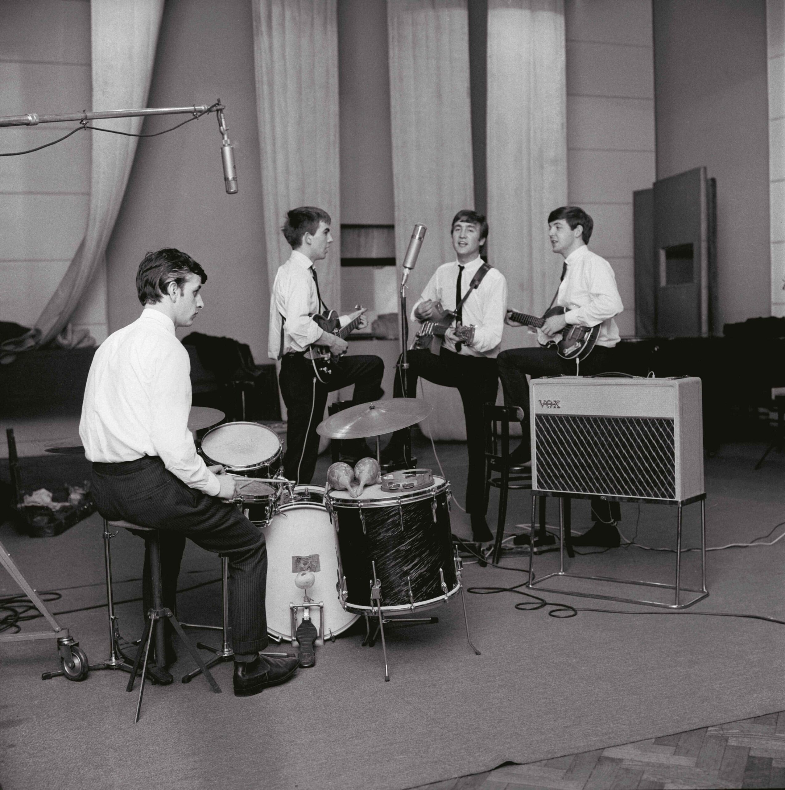 A black-and-white photograph shows The Beatles during a recording session at Abbey Road Studios in London.  The Band Members The four members are dressed uniformly in white button-down shirts and thin dark ties:  Ringo Starr: In the foreground on the left, seated at his drum kit. A pair of maracas and a tambourine rest on the floor tom in front of him.  George Harrison: Standing in profile to the right of Ringo, holding an electric guitar.  John Lennon: Positioned in the center, singing into a tall condenser microphone while playing an acoustic-electric guitar.  Paul McCartney: On the right, playing his signature Höfner "violin" bass and looking toward John.  Studio Environment Equipment: A large Vox amplifier on a chrome stand is visible in the right foreground. A boom microphone hangs over the drums from the top left corner.  Background: The studio features high ceilings with long, vertical acoustic baffles or curtains hanging in the background. The floor is covered by a large neutral-colored rug over a parquet wood floor.  Atmosphere: The composition captures a professional yet candid moment of the band working together early in their career.