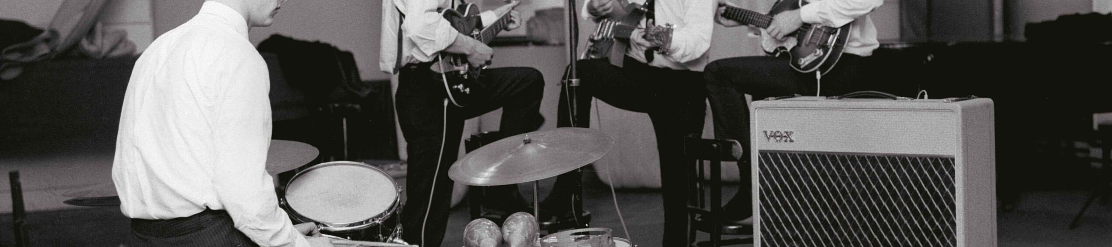 A black-and-white photograph shows The Beatles during a recording session at Abbey Road Studios in London. The Band Members The four members are dressed uniformly in white button-down shirts and thin dark ties: Ringo Starr: In the foreground on the left, seated at his drum kit. A pair of maracas and a tambourine rest on the floor tom in front of him. George Harrison: Standing in profile to the right of Ringo, holding an electric guitar. John Lennon: Positioned in the center, singing into a tall condenser microphone while playing an acoustic-electric guitar. Paul McCartney: On the right, playing his signature Höfner "violin" bass and looking toward John. Studio Environment Equipment: A large Vox amplifier on a chrome stand is visible in the right foreground. A boom microphone hangs over the drums from the top left corner. Background: The studio features high ceilings with long, vertical acoustic baffles or curtains hanging in the background. The floor is covered by a large neutral-colored rug over a parquet wood floor. Atmosphere: The composition captures a professional yet candid moment of the band working together early in their career.