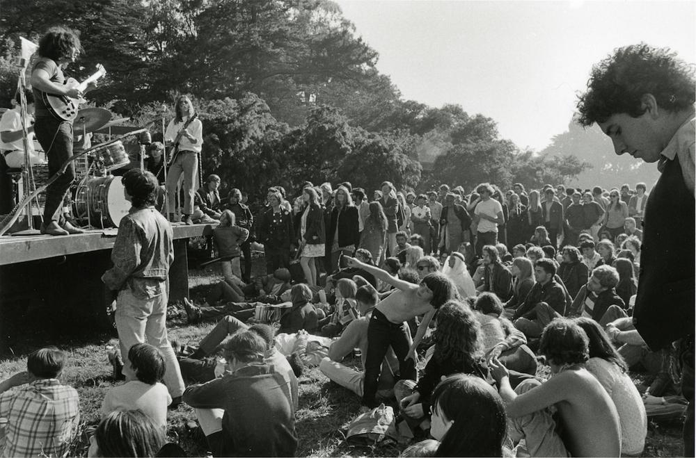 A candid black-and-white photograph shows a band performing on a makeshift outdoor stage, likely a flatbed trailer, in a park setting. On the left, a guitarist with long, curly dark hair plays an electric guitar while other band members perform behind him. In front of the stage, a large crowd of young people sits and reclines on the grass, watching the concert. In the immediate right foreground, a man is visible in profile, looking toward the stage. The background is filled with lush trees under bright, natural light.
