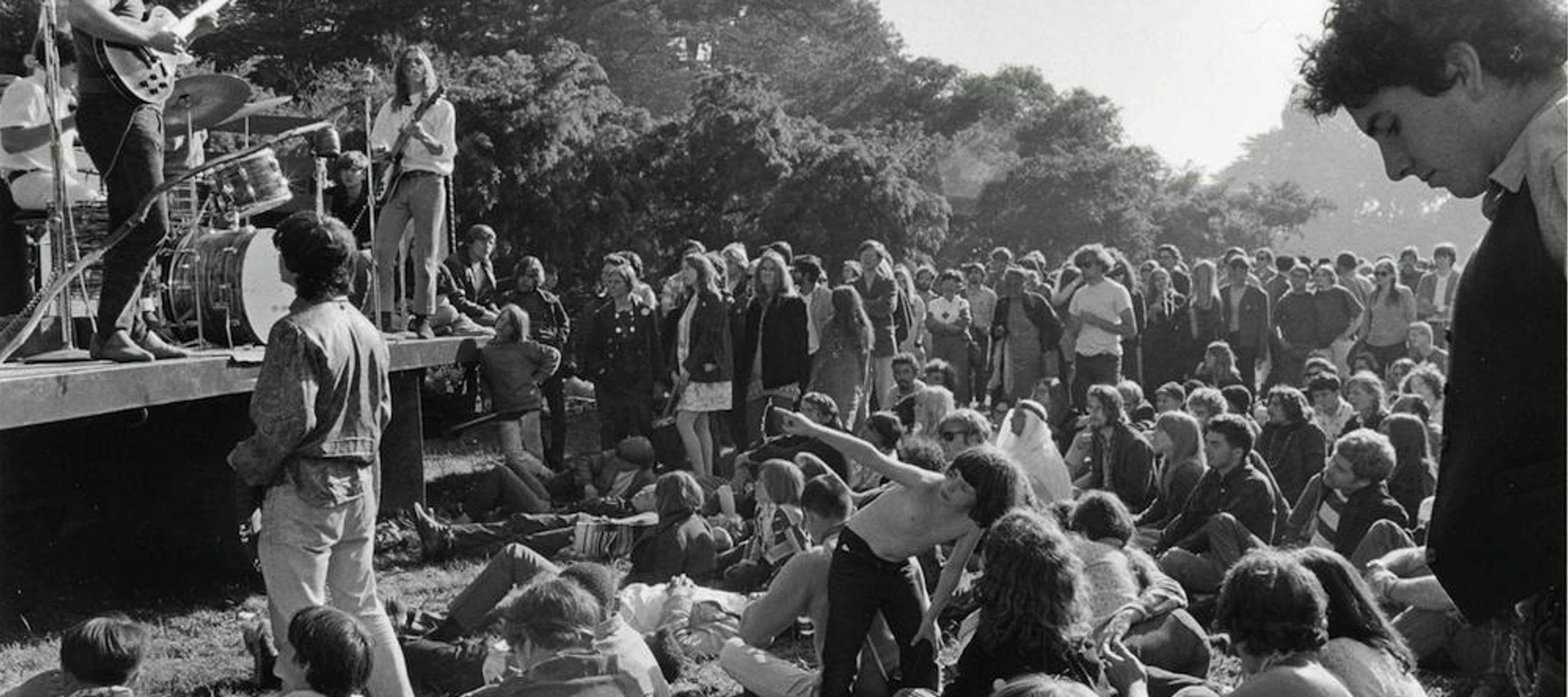 A candid black-and-white photograph shows a band performing on a makeshift outdoor stage, likely a flatbed trailer, in a park setting. On the left, a guitarist with long, curly dark hair plays an electric guitar while other band members perform behind him. In front of the stage, a large crowd of young people sits and reclines on the grass, watching the concert. In the immediate right foreground, a man is visible in profile, looking toward the stage. The background is filled with lush trees under bright, natural light.