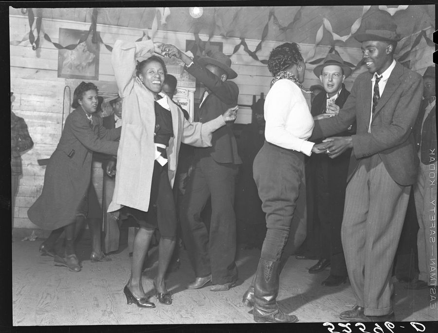 A candid, black-and-white photograph capturing a lively social gathering where people are dancing indoors. In the foreground, a woman wearing a light-colored coat over a dark dress dances with her arms raised. Beside her, a man wearing a suit, tie, and fedora dances with a woman dressed in dark trousers and tall boots. The background shows wood-paneled walls decorated with streamers, and other guests watching or participating in the festivities.