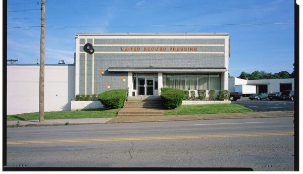 This image shows the exterior of the United Record Pressing facility.  The Building: It is a mid-century, one-story commercial building featuring gray, patterned brickwork and the company name, "UNITED RECORD PRESSING," displayed in large, dark lettering near the roofline. A logo featuring two vinyl records is visible on the upper left side.