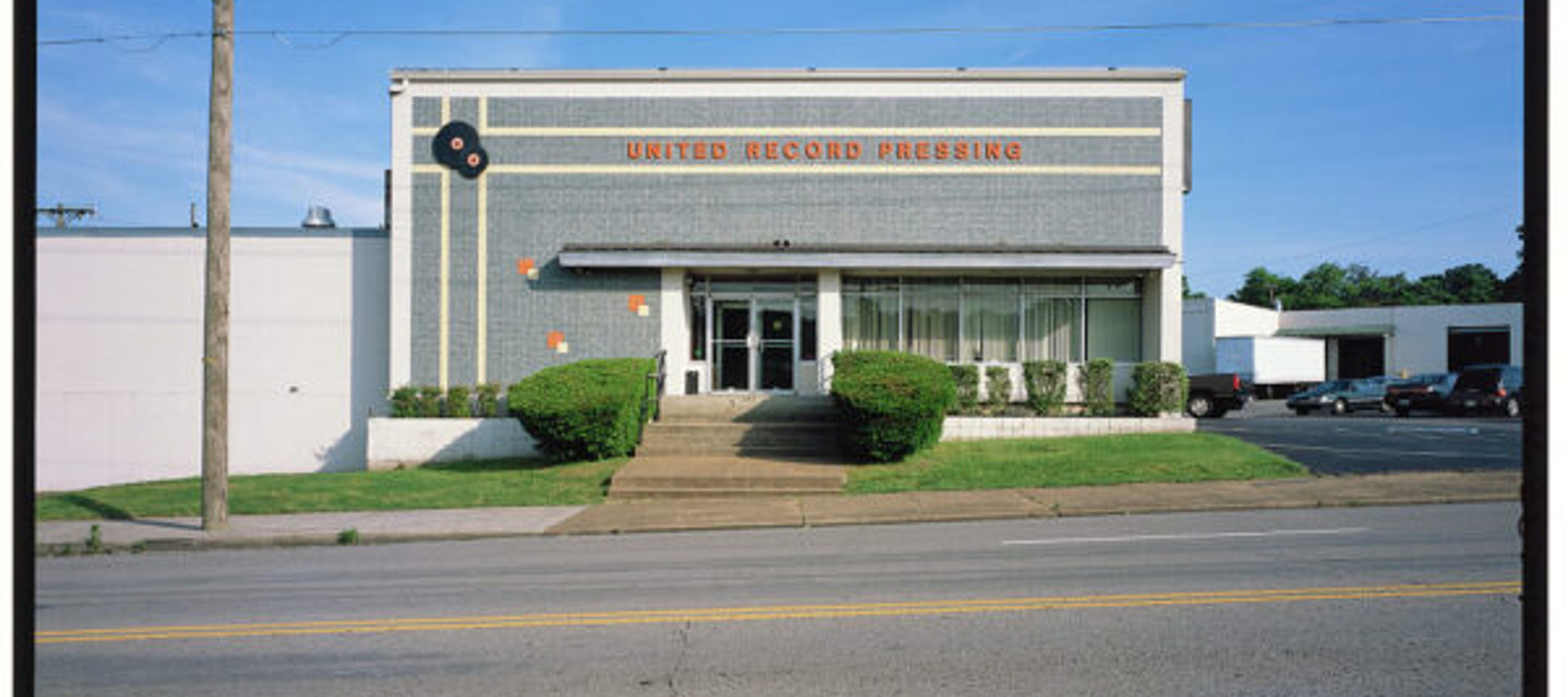 This image shows the exterior of the United Record Pressing facility. The Building: It is a mid-century, one-story commercial building featuring gray, patterned brickwork and the company name, "UNITED RECORD PRESSING," displayed in large, dark lettering near the roofline. A logo featuring two vinyl records is visible on the upper left side.