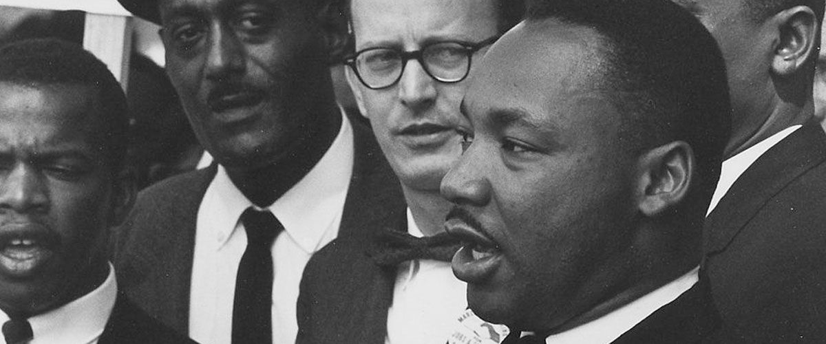 A black and white close-up photograph features Dr. Martin Luther King Jr. in profile on the right, speaking with his mouth open. He is surrounded by several other men in suits and ties, including John Lewis on the far left. The men appear to be participating in a serious event, likely a civil rights march or protest, as they look forward with focused expressions.
