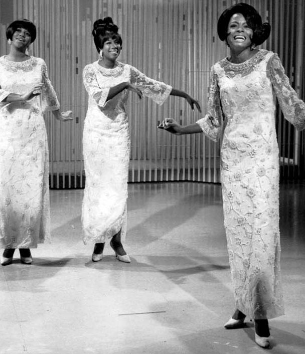 A black and white photo of the Supremes performing in white dresses.
