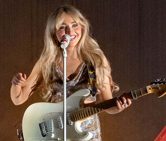 Sabrina Carpenter smiling behind a microphone with a guitar