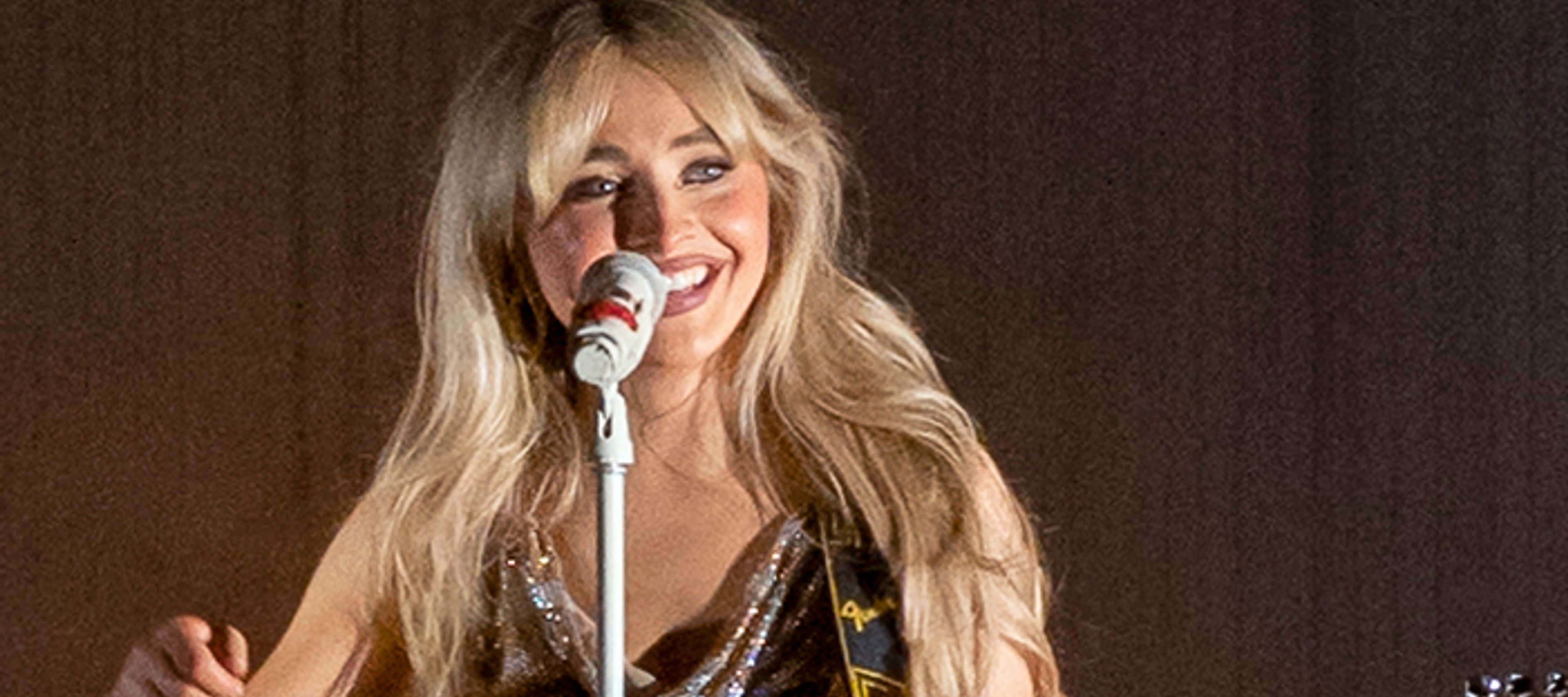 Sabrina Carpenter smiling behind a microphone with a guitar
