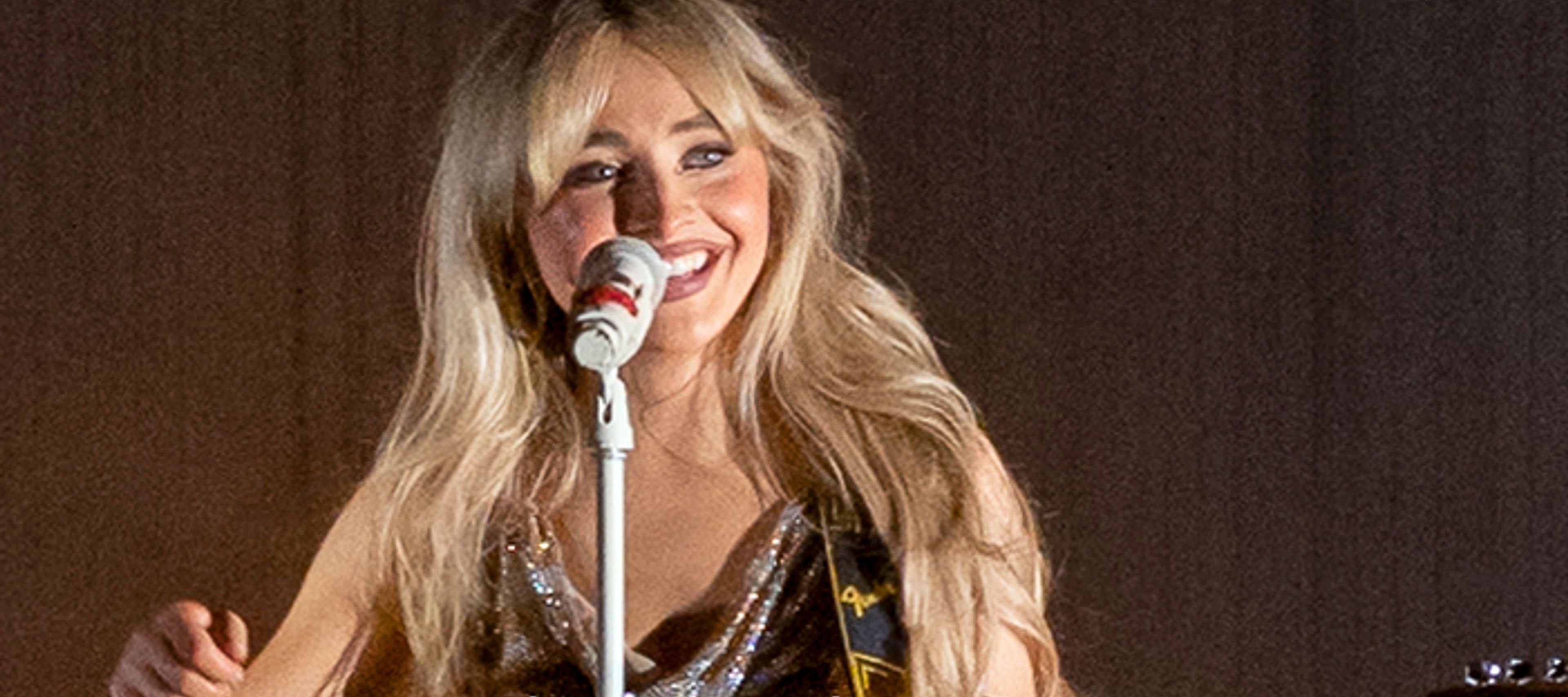 Sabrina Carpenter smiling behind a microphone with a guitar