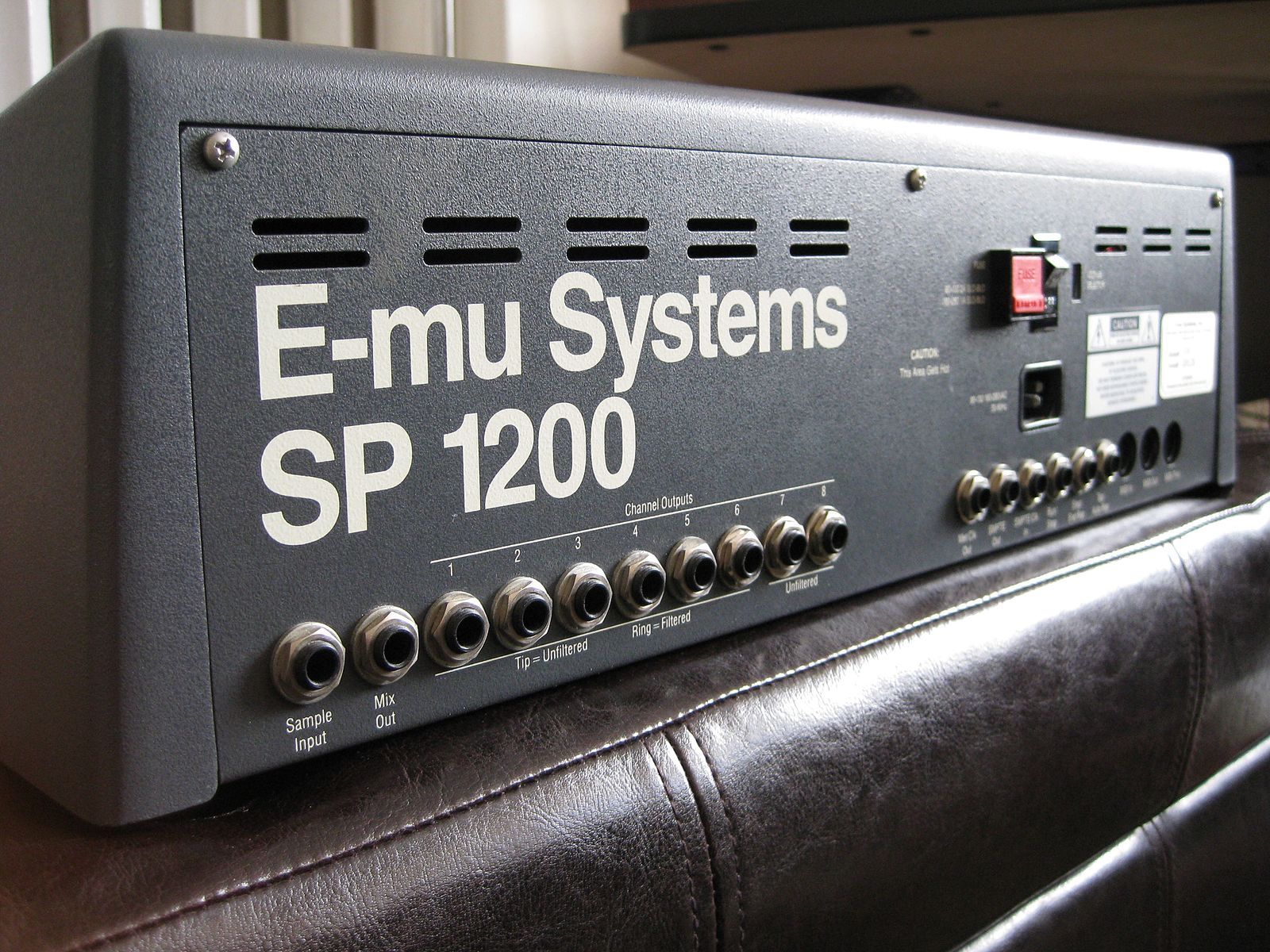A close-up, angled shot of the rear panel of a vintage E-mu Systems SP-1200 drum machine and sampler. The device is housed in a dark grey, textured metal casing with large white lettering that reads "E-mu Systems SP 1200." A row of 1/4-inch audio jacks is visible along the bottom, labeled for "Sample Input," "Mix Out," and eight individual "Channel Outputs." The unit is resting on a dark brown leather surface.