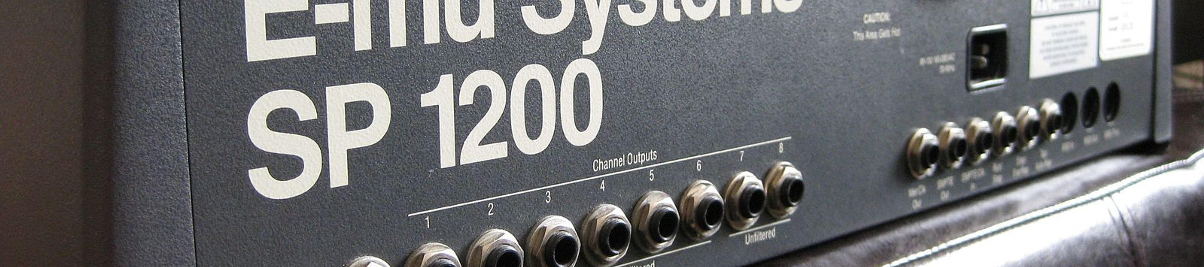 A close-up, angled shot of the rear panel of a vintage E-mu Systems SP-1200 drum machine and sampler. The device is housed in a dark grey, textured metal casing with large white lettering that reads "E-mu Systems SP 1200." A row of 1/4-inch audio jacks is visible along the bottom, labeled for "Sample Input," "Mix Out," and eight individual "Channel Outputs." The unit is resting on a dark brown leather surface.