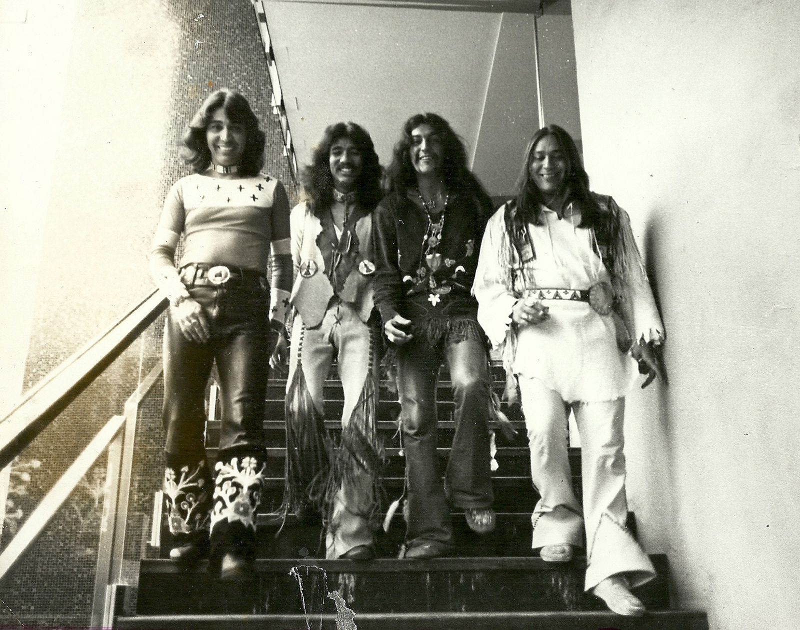 A black-and-white, portrait-oriented photograph captures four members of the Native American rock band Redbone walking down an outdoor staircase, smiling warmly.  The men are lined up side-by-side, dressed in eclectic, 1970s style that blends contemporary rock fashion with traditional Indigenous clothing elements:  The man on the far left wears a fitted, long-sleeved shirt patterned with small crosses across the chest, dark leather trousers accented with highly detailed, embroidered floral patterns on the flared cuffs, and a thick belt with a large buckle.  The second man from the left wears a light-colored vest with fringed edges over a bare chest, accessorized with a choker and a long pendant. He wears matching light-colored trousers heavily decorated with long fringe draping down the sides of the legs.  The third man from the left wears a dark, patterned long-sleeved shirt, layered necklaces, and leather trousers featuring long fringe around the waist and hips.  The man on the far right wears a light-colored, fringed tunic with a patterned belt and matching light-colored trousers.  They are descending dark stairs positioned between a textured brick wall on the left and a smooth, light-colored wall on the right. A metal handrail runs down the left side of the staircase.