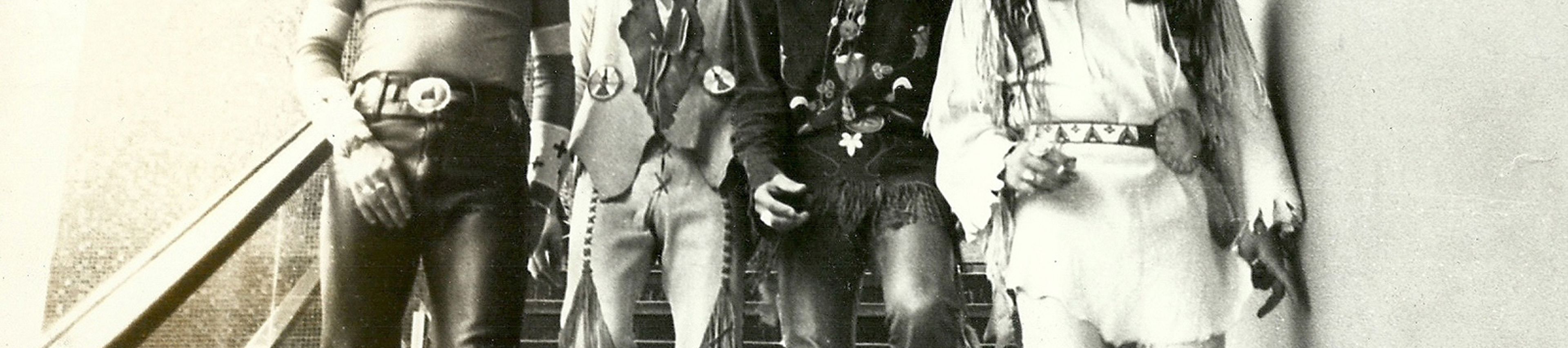 A black-and-white, portrait-oriented photograph captures four members of the Native American rock band Redbone walking down an outdoor staircase, smiling warmly. The men are lined up side-by-side, dressed in eclectic, 1970s style that blends contemporary rock fashion with traditional Indigenous clothing elements: The man on the far left wears a fitted, long-sleeved shirt patterned with small crosses across the chest, dark leather trousers accented with highly detailed, embroidered floral patterns on the flared cuffs, and a thick belt with a large buckle. The second man from the left wears a light-colored vest with fringed edges over a bare chest, accessorized with a choker and a long pendant. He wears matching light-colored trousers heavily decorated with long fringe draping down the sides of the legs. The third man from the left wears a dark, patterned long-sleeved shirt, layered necklaces, and leather trousers featuring long fringe around the waist and hips. The man on the far right wears a light-colored, fringed tunic with a patterned belt and matching light-colored trousers. They are descending dark stairs positioned between a textured brick wall on the left and a smooth, light-colored wall on the right. A metal handrail runs down the left side of the staircase.