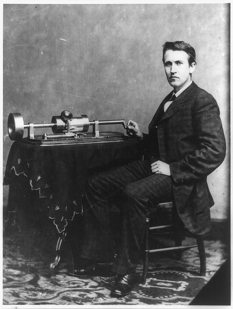 A black and white image of a young Thomas Edison, seated in a suit, with a hand on his phonograph machine, which sits on a table with a tablecloth.