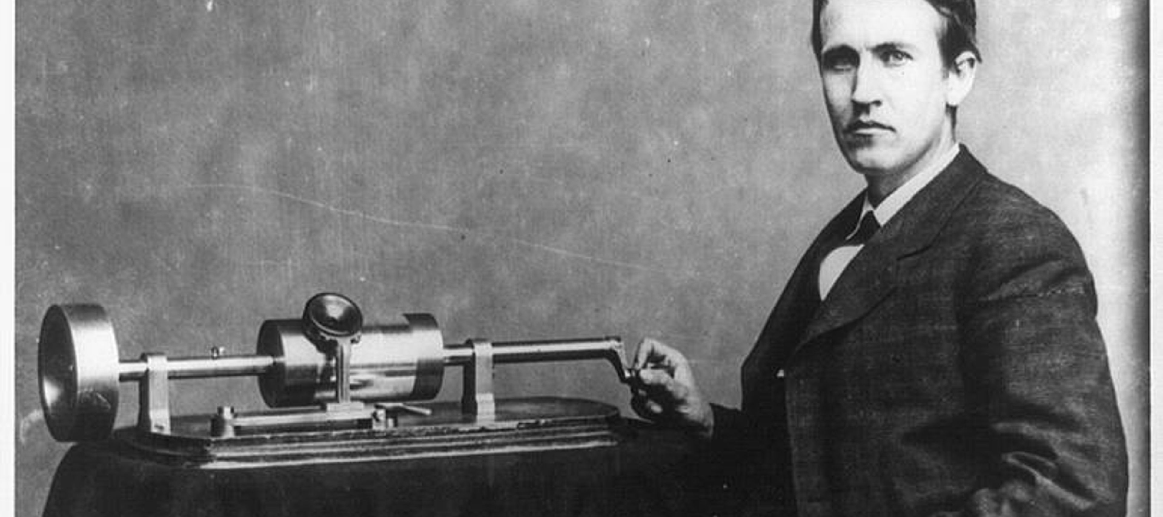A black and white image of a young Thomas Edison, seated in a suit, with a hand on his phonograph machine, which sits on a table with a tablecloth.