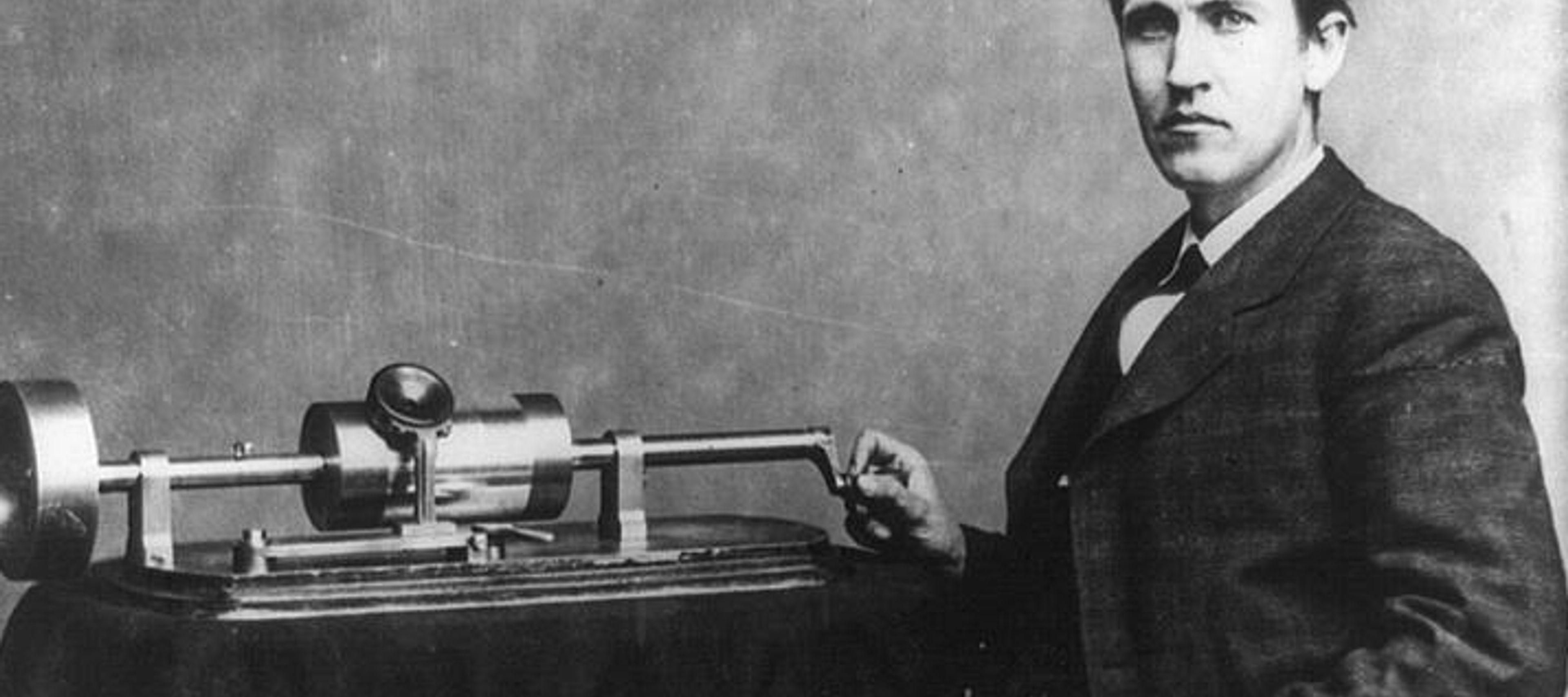 A black and white image of a young Thomas Edison, seated in a suit, with a hand on his phonograph machine, which sits on a table with a tablecloth.