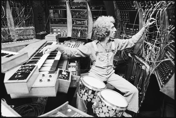 A black-and-white photograph of a man with curly hair, wearing overalls, seated in a recording studio surrounded by a massive, complex modular synthesizer labeled "TONTO." He is actively adjusting patch cables on the synthesizer while his other hand rests on a keyboard. Two small drums are visible in the foreground.