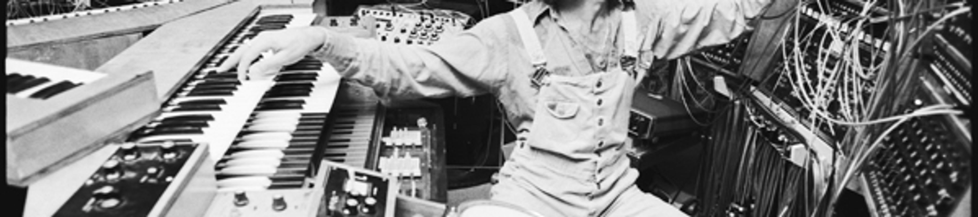 A black-and-white photograph of a man with curly hair, wearing overalls, seated in a recording studio surrounded by a massive, complex modular synthesizer labeled "TONTO." He is actively adjusting patch cables on the synthesizer while his other hand rests on a keyboard. Two small drums are visible in the foreground.