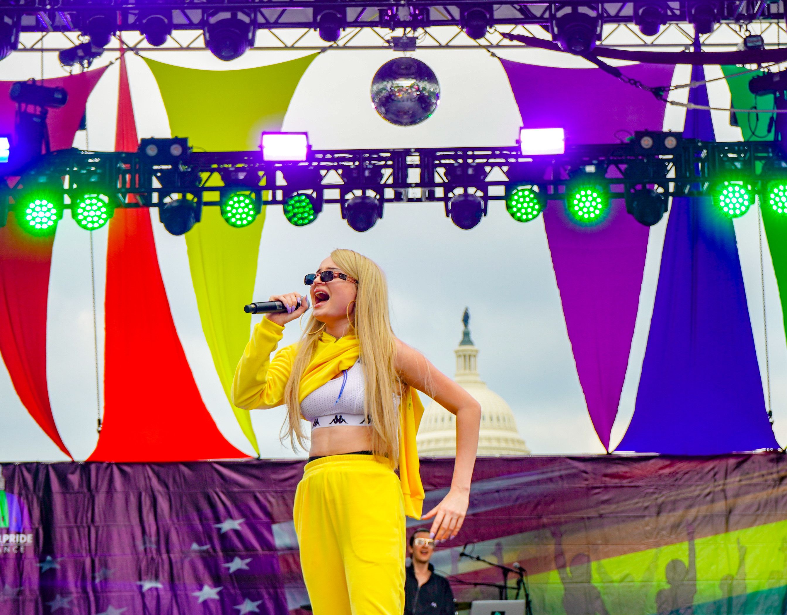 Kim Petras signing on a stage with rainbow decorations and a disco ball, in front of the US Capitol building