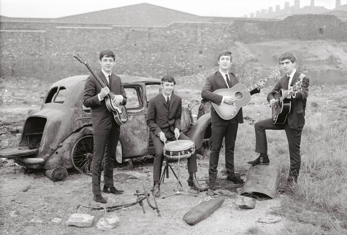 A black-and-white photograph shows The Beatles posing with their instruments in a bleak, derelict landscape in Liverpool.  The Band Members The four members are dressed in matching dark, slim-fitting suits and ties:  Paul McCartney: On the far left, standing beside a rusted car and holding his Höfner "violin" bass.  Ringo Starr: Seated in the center on a small stool, smiling while holding drumsticks over a single snare drum.  John Lennon: To the right of Ringo, standing and holding an acoustic-electric guitar.  George Harrison: On the far right, standing with one foot propped up on a piece of debris while holding a dark electric guitar.  Setting and Background The Environment: The band is positioned in a wasteland or "bombsite" filled with dirt, sparse grass, and scattered debris.  The Car: A heavily rusted, windowless vintage car shell sits behind Paul and Ringo, with its trunk open and wheels missing.  The Skyline: In the background, a long stone wall or embankment stretches across the frame, with the faint silhouettes of distant, repetitive rooftops (likely terraced housing) visible under a pale, overcast sky.  The image captures a juxtaposition between the band's clean, professional appearance and the gritty, post-war urban decay of their hometown.
