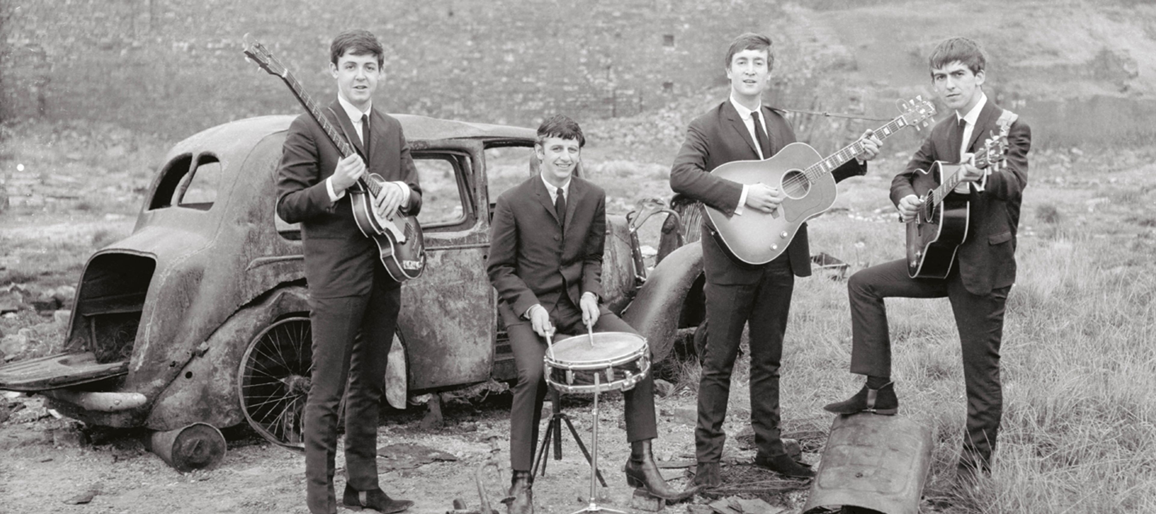A black-and-white photograph shows The Beatles posing with their instruments in a bleak, derelict landscape in Liverpool. The Band Members The four members are dressed in matching dark, slim-fitting suits and ties: Paul McCartney: On the far left, standing beside a rusted car and holding his Höfner "violin" bass. Ringo Starr: Seated in the center on a small stool, smiling while holding drumsticks over a single snare drum. John Lennon: To the right of Ringo, standing and holding an acoustic-electric guitar. George Harrison: On the far right, standing with one foot propped up on a piece of debris while holding a dark electric guitar. Setting and Background The Environment: The band is positioned in a wasteland or "bombsite" filled with dirt, sparse grass, and scattered debris. The Car: A heavily rusted, windowless vintage car shell sits behind Paul and Ringo, with its trunk open and wheels missing. The Skyline: In the background, a long stone wall or embankment stretches across the frame, with the faint silhouettes of distant, repetitive rooftops (likely terraced housing) visible under a pale, overcast sky. The image captures a juxtaposition between the band's clean, professional appearance and the gritty, post-war urban decay of their hometown.