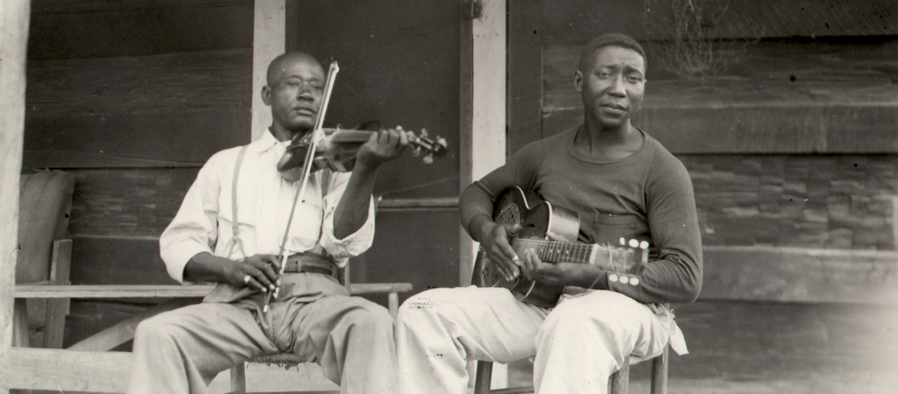 A black-and-white, landscape-oriented photograph shows two Black men seated on a porch, playing instruments. The man on the left wears a light-colored, long-sleeved button-down shirt with suspenders and light-colored trousers; he is playing a violin. The man on the right wears a dark, long-sleeved shirt and light-colored trousers; he is playing an acoustic guitar. They are both seated on simple wooden chairs in front of a rustic wooden building.