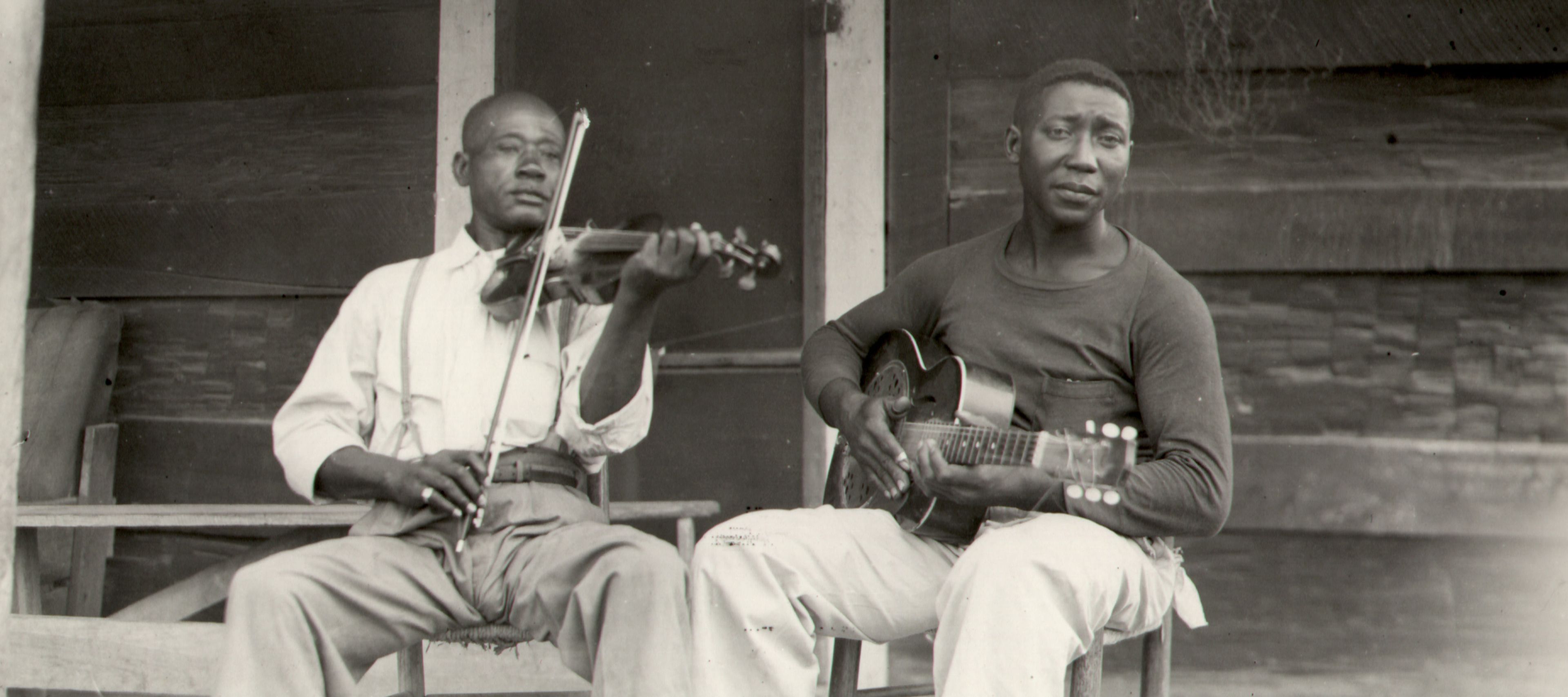 A black-and-white, landscape-oriented photograph shows two Black men seated on a porch, playing instruments. The man on the left wears a light-colored, long-sleeved button-down shirt with suspenders and light-colored trousers; he is playing a violin. The man on the right wears a dark, long-sleeved shirt and light-colored trousers; he is playing an acoustic guitar. They are both seated on simple wooden chairs in front of a rustic wooden building.