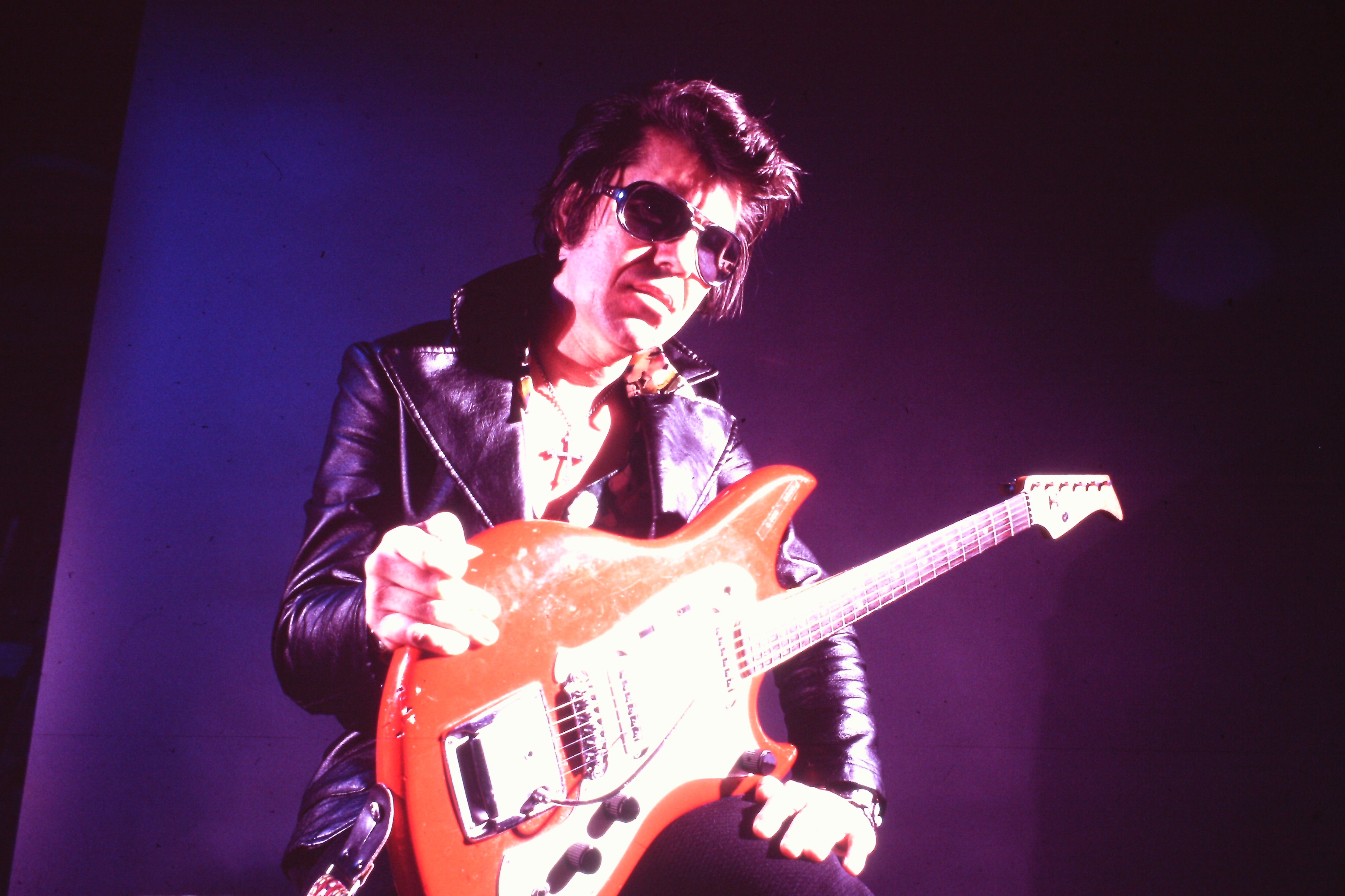 A landscape-oriented, low-angle photograph, bathed in dramatic purple stage lighting, captures Shawnee guitarist Link Wray posing with an electric guitar.  Wray is positioned in the center of the frame, looking down toward the camera. He has dark, swept-back hair and wears dark sunglasses, a black leather jacket with wide lapels over a patterned shirt, and a cross pendant necklace.  He holds a bright red electric guitar across his lap, resting his right hand near the top of the body and his left hand near the bottom. The background is a solid, dark purple wall illuminated by a strong spotlight casting a bright glow on the right side of Wray's face and his guitar.