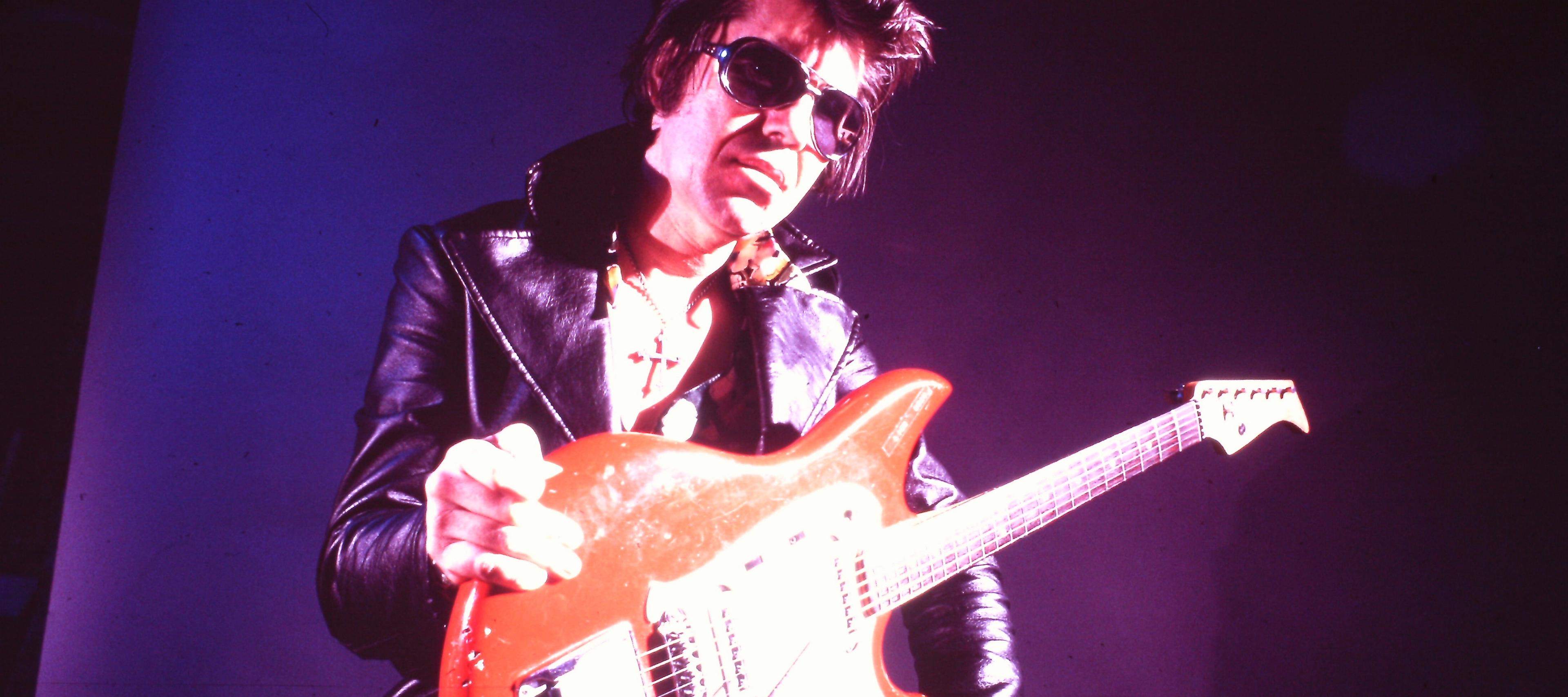 A landscape-oriented, low-angle photograph, bathed in dramatic purple stage lighting, captures Shawnee guitarist Link Wray posing with an electric guitar. Wray is positioned in the center of the frame, looking down toward the camera. He has dark, swept-back hair and wears dark sunglasses, a black leather jacket with wide lapels over a patterned shirt, and a cross pendant necklace. He holds a bright red electric guitar across his lap, resting his right hand near the top of the body and his left hand near the bottom. The background is a solid, dark purple wall illuminated by a strong spotlight casting a bright glow on the right side of Wray's face and his guitar.