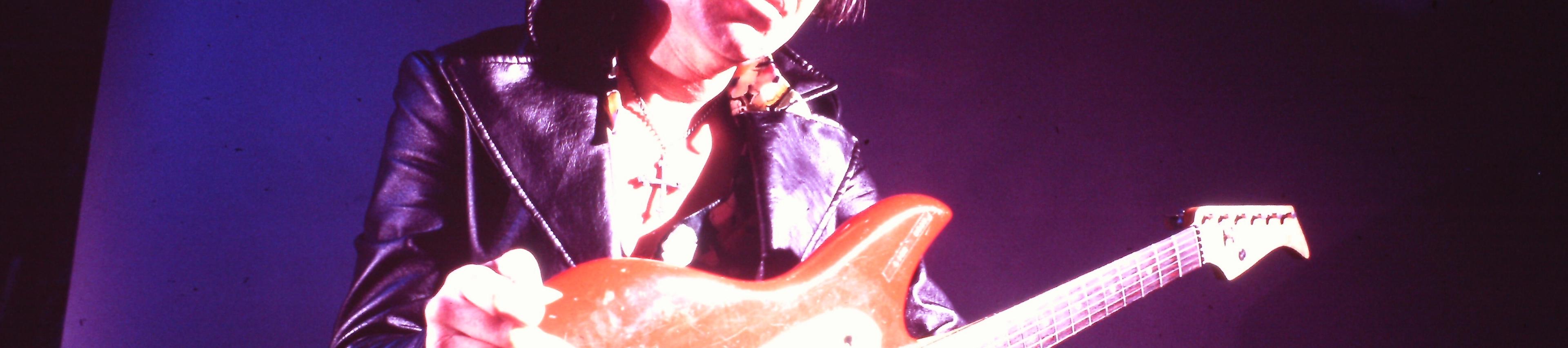 A landscape-oriented, low-angle photograph, bathed in dramatic purple stage lighting, captures Shawnee guitarist Link Wray posing with an electric guitar. Wray is positioned in the center of the frame, looking down toward the camera. He has dark, swept-back hair and wears dark sunglasses, a black leather jacket with wide lapels over a patterned shirt, and a cross pendant necklace. He holds a bright red electric guitar across his lap, resting his right hand near the top of the body and his left hand near the bottom. The background is a solid, dark purple wall illuminated by a strong spotlight casting a bright glow on the right side of Wray's face and his guitar.