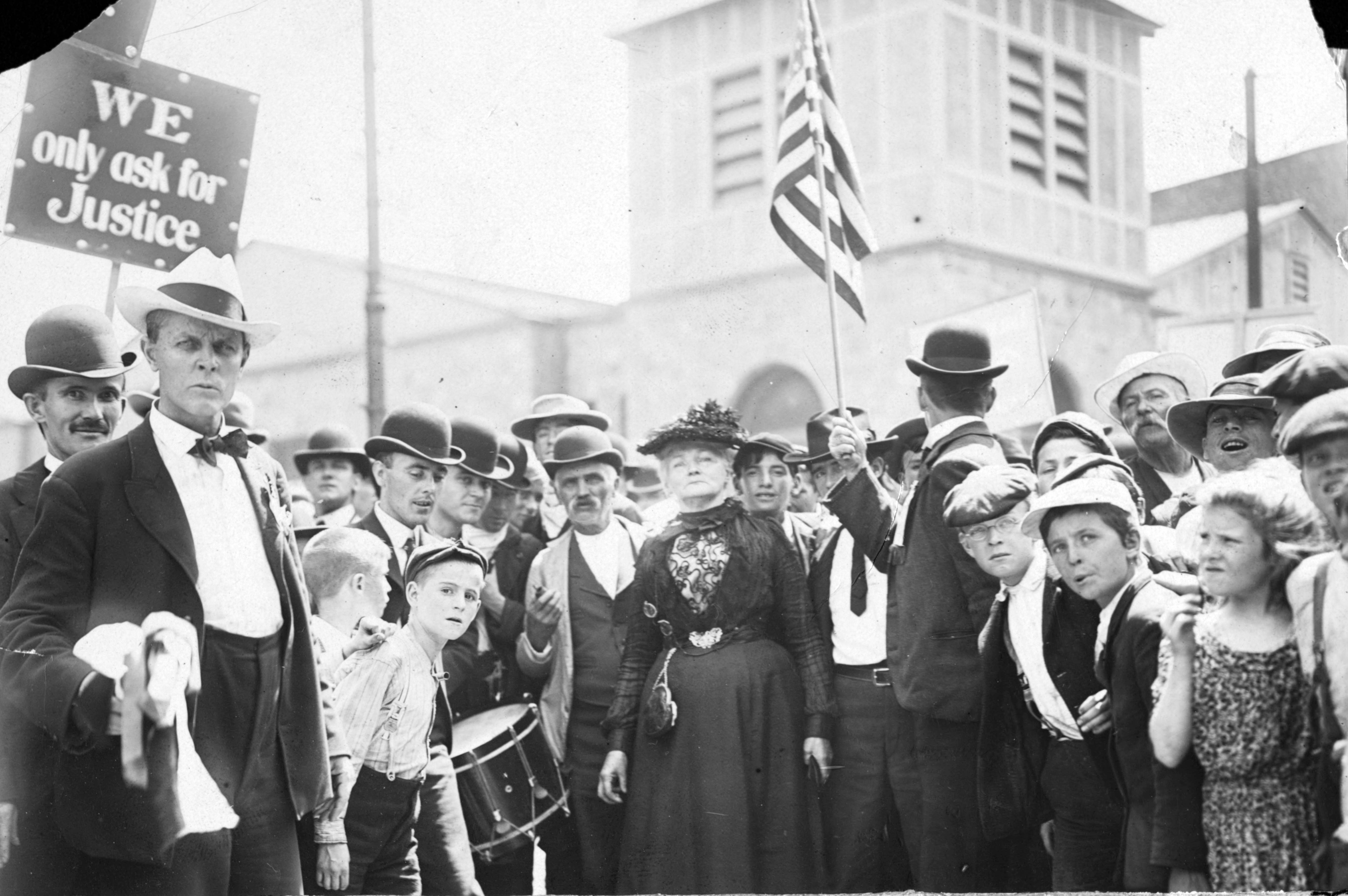 A black-and-white historical photograph depicts a large crowd of people gathered outdoors for a protest or march, likely in the early 20th century.  In the foreground, a man in a light-colored fedora and a dark suit stands prominently on the left, looking directly at the camera with a serious expression. To his right, a young boy with a drum and a woman in a formal, dark Victorian-style dress and feathered hat are visible among the densely packed crowd. Several men in the group wear bowler hats, and a few children are scattered throughout.  In the upper left corner, a sign is held aloft that reads, "WE only ask for Justice." To the right, another person holds an American flag. The background shows the blurred architecture of buildings, suggesting an urban or town square setting. The overall mood is solemn and determined.
