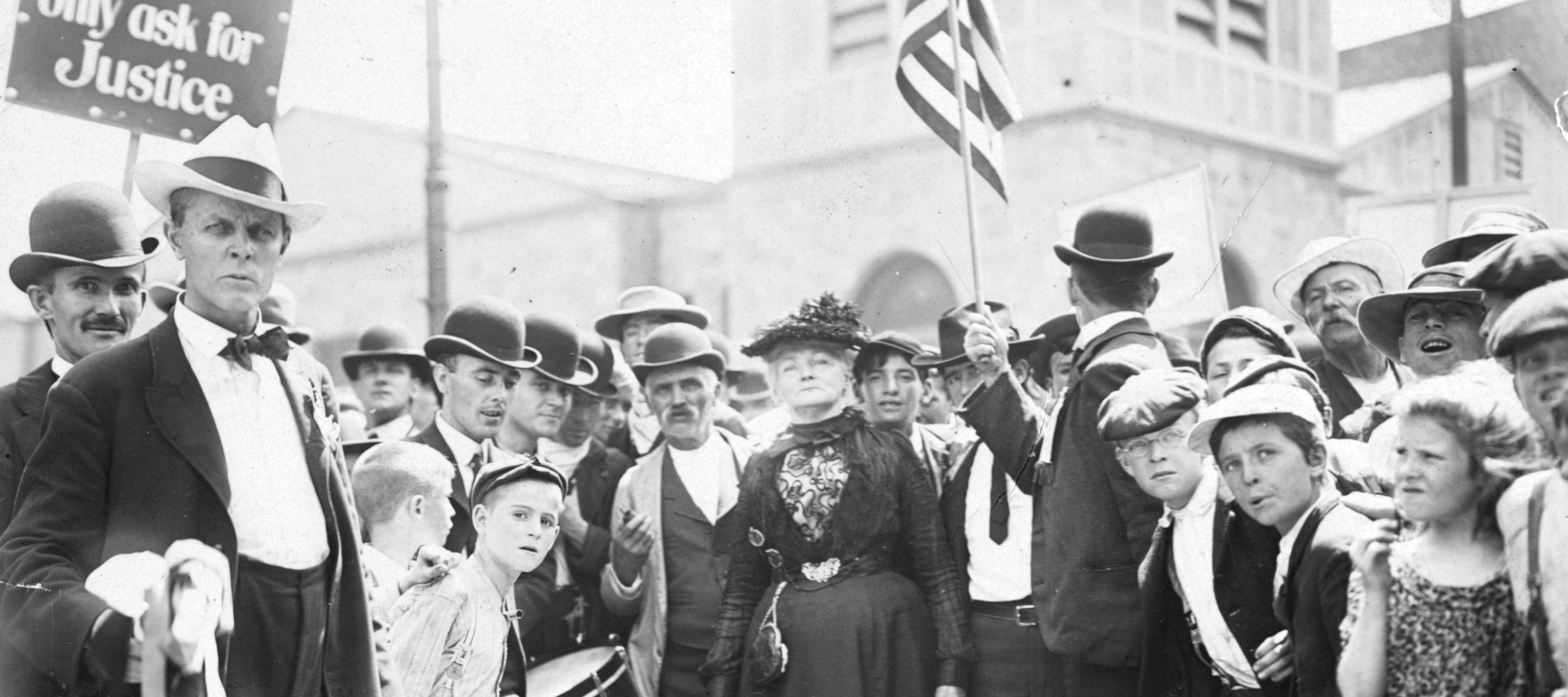 A black-and-white historical photograph depicts a large crowd of people gathered outdoors for a protest or march, likely in the early 20th century. In the foreground, a man in a light-colored fedora and a dark suit stands prominently on the left, looking directly at the camera with a serious expression. To his right, a young boy with a drum and a woman in a formal, dark Victorian-style dress and feathered hat are visible among the densely packed crowd. Several men in the group wear bowler hats, and a few children are scattered throughout. In the upper left corner, a sign is held aloft that reads, "WE only ask for Justice." To the right, another person holds an American flag. The background shows the blurred architecture of buildings, suggesting an urban or town square setting. The overall mood is solemn and determined.