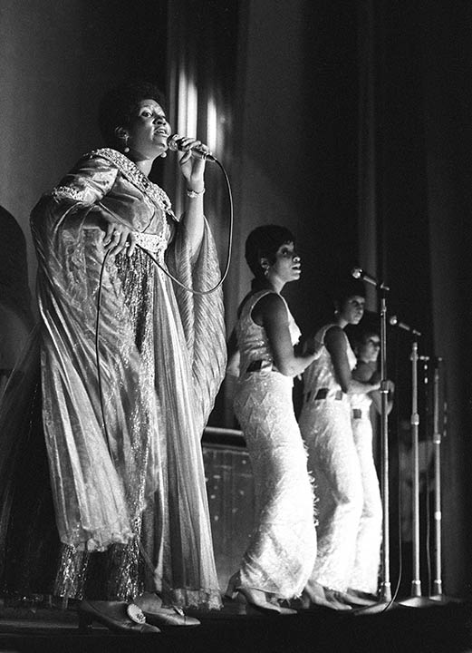 A black and white photograph captures Aretha Franklin performing on stage, bathed in a spotlight. She stands on the left, singing into a wired microphone and wearing a shimmering, floor-length gown with flowing, cape-like sleeves. To her right, three backup singers in matching, sleeveless sequined dresses stand in a diagonal line, each positioned behind their own microphone. The stage is dark, emphasizing the performers and the glitter of their stage attire.