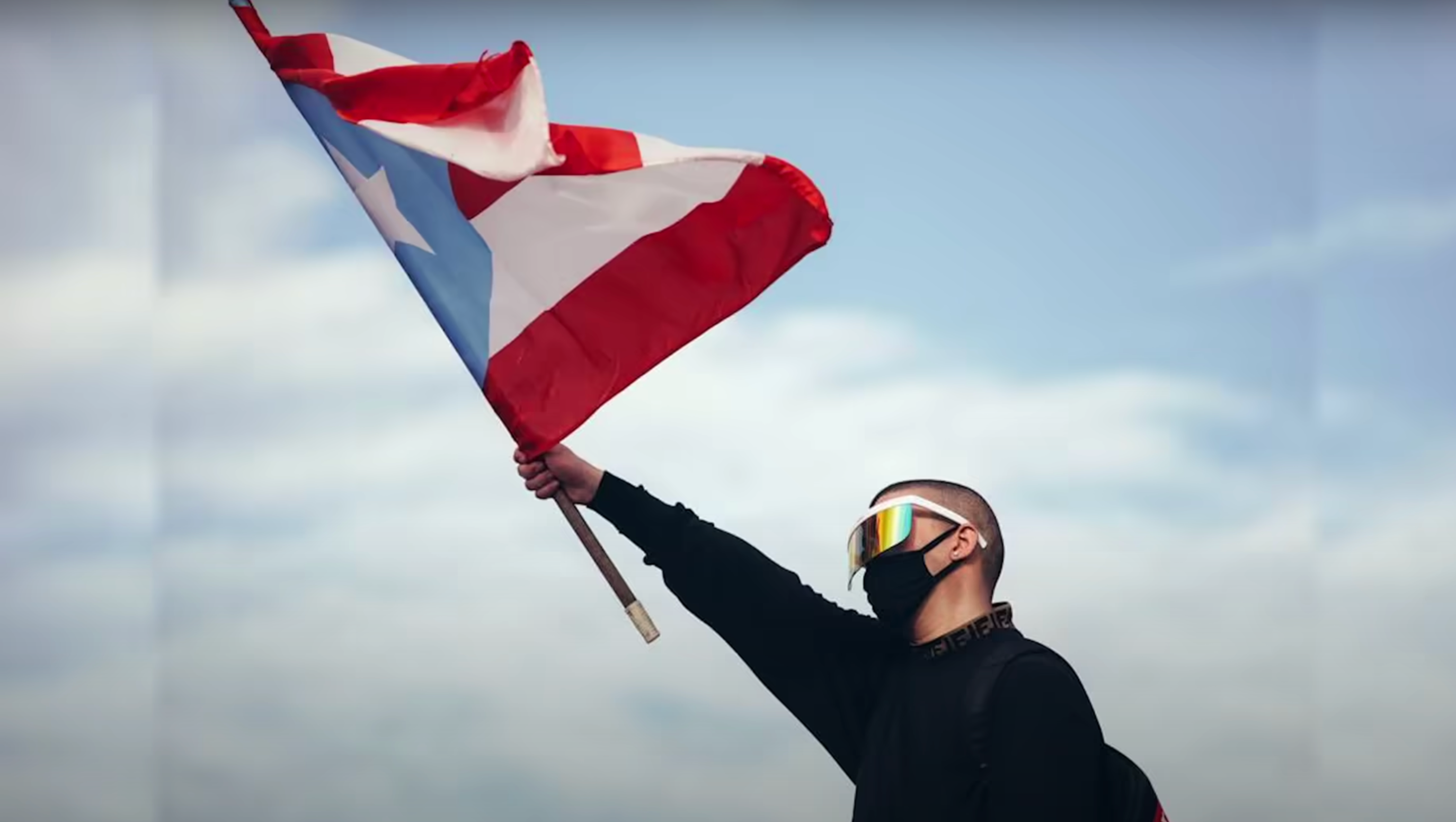 A screen capture of the Bad Bunny video "El Apagón" featuring Bad Bunny wearing a visor and mask waving the red, white, and blue Puerto Rican Flag