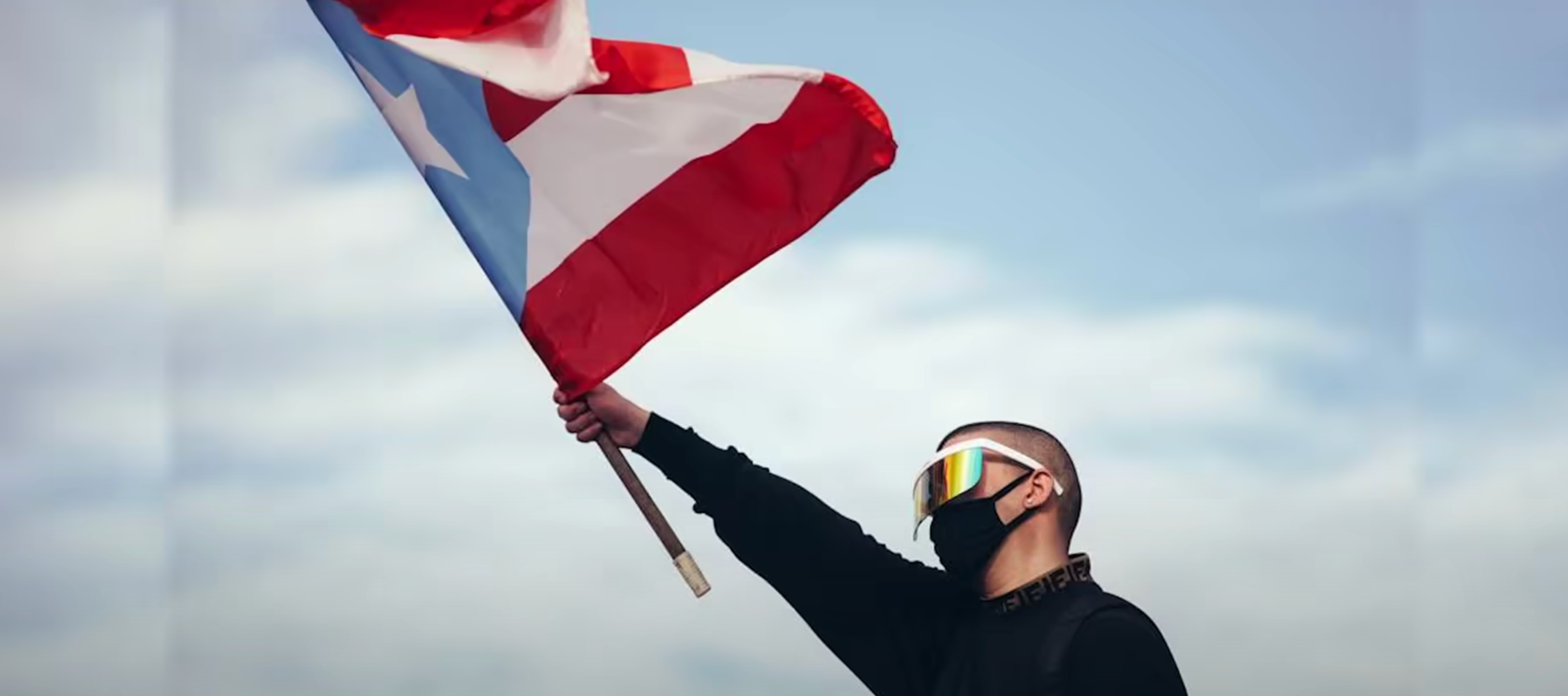 A screen capture of the Bad Bunny video "El Apagón" featuring Bad Bunny wearing a visor and mask waving the red, white, and blue Puerto Rican Flag