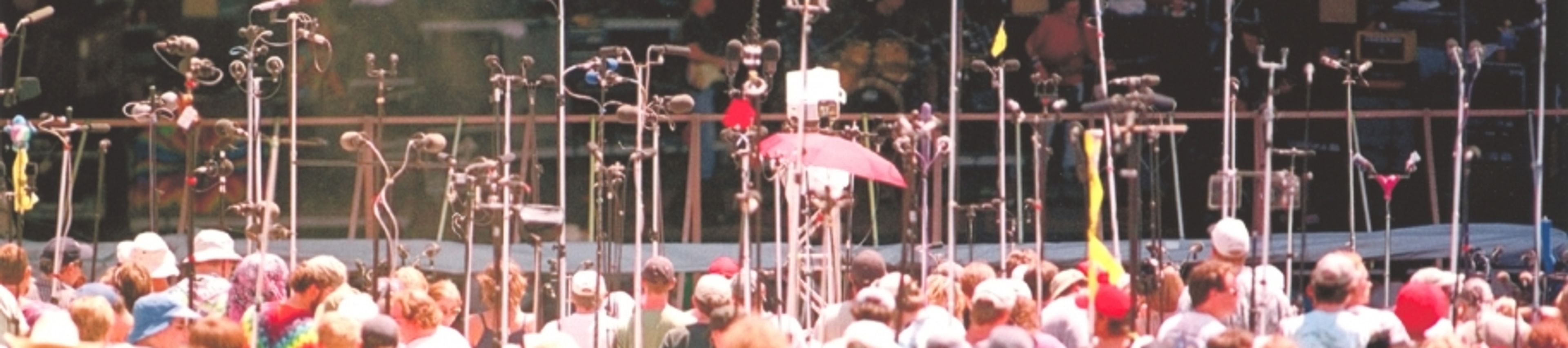 A wide, outdoor concert scene featuring a dense, overwhelming cluster of microphone stands and boom arms in the foreground that partially obstruct the view of the stage. Behind this "forest" of microphones, band members are visible on stage, with a large crowd gathered in front of them under the bright sun, typical of an outdoor music festival.