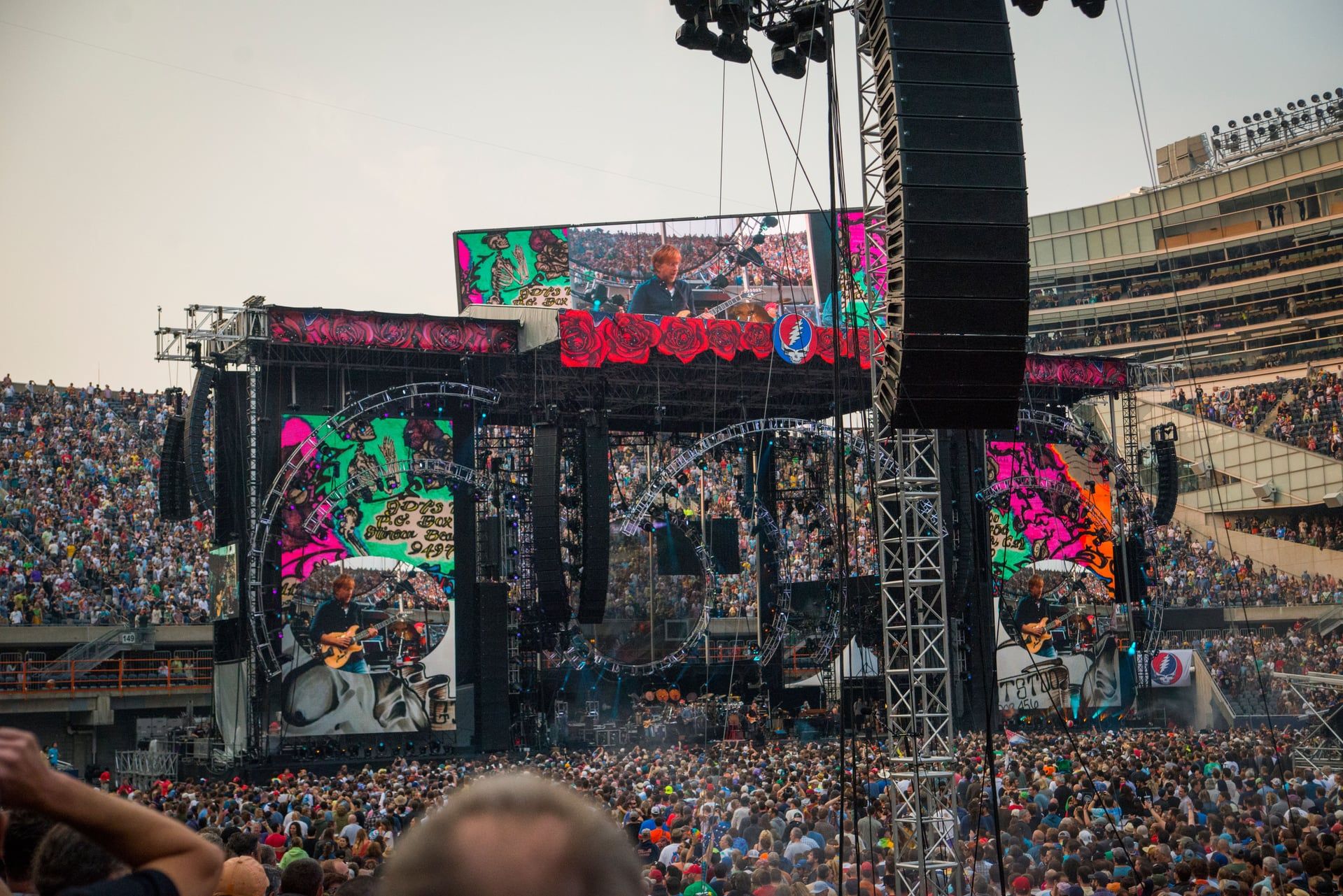 A wide-angle view of a large outdoor stadium concert filled with a massive crowd of fans. The stage is elaborate, featuring towering speaker arrays and large LED screens that display live footage of a musician playing guitar. The iconic Grateful Dead "Steal Your Face" skull logo is prominently displayed on the stage rigging. The scene is captured from the perspective of the audience, with concert equipment visible in the foreground under an open sky.