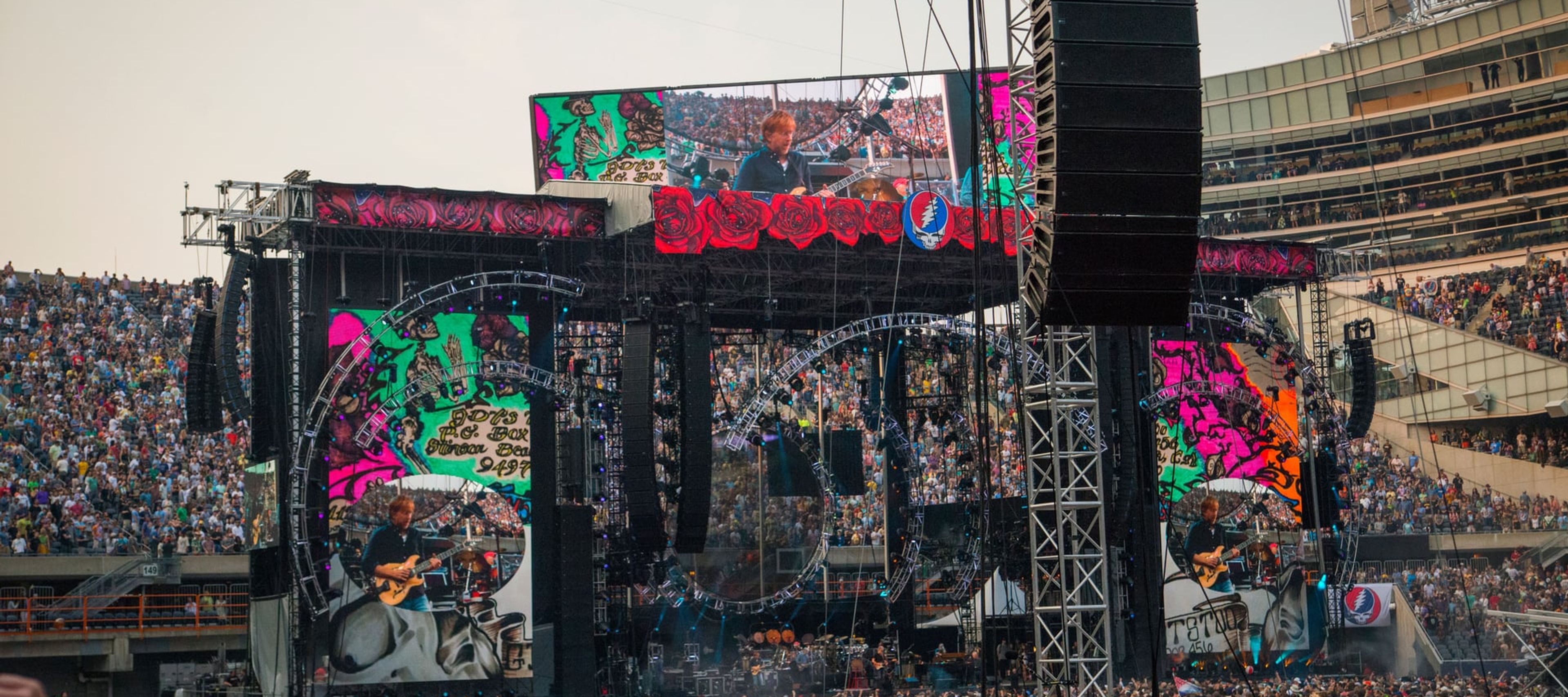 A wide-angle view of a large outdoor stadium concert filled with a massive crowd of fans. The stage is elaborate, featuring towering speaker arrays and large LED screens that display live footage of a musician playing guitar. The iconic Grateful Dead "Steal Your Face" skull logo is prominently displayed on the stage rigging. The scene is captured from the perspective of the audience, with concert equipment visible in the foreground under an open sky.