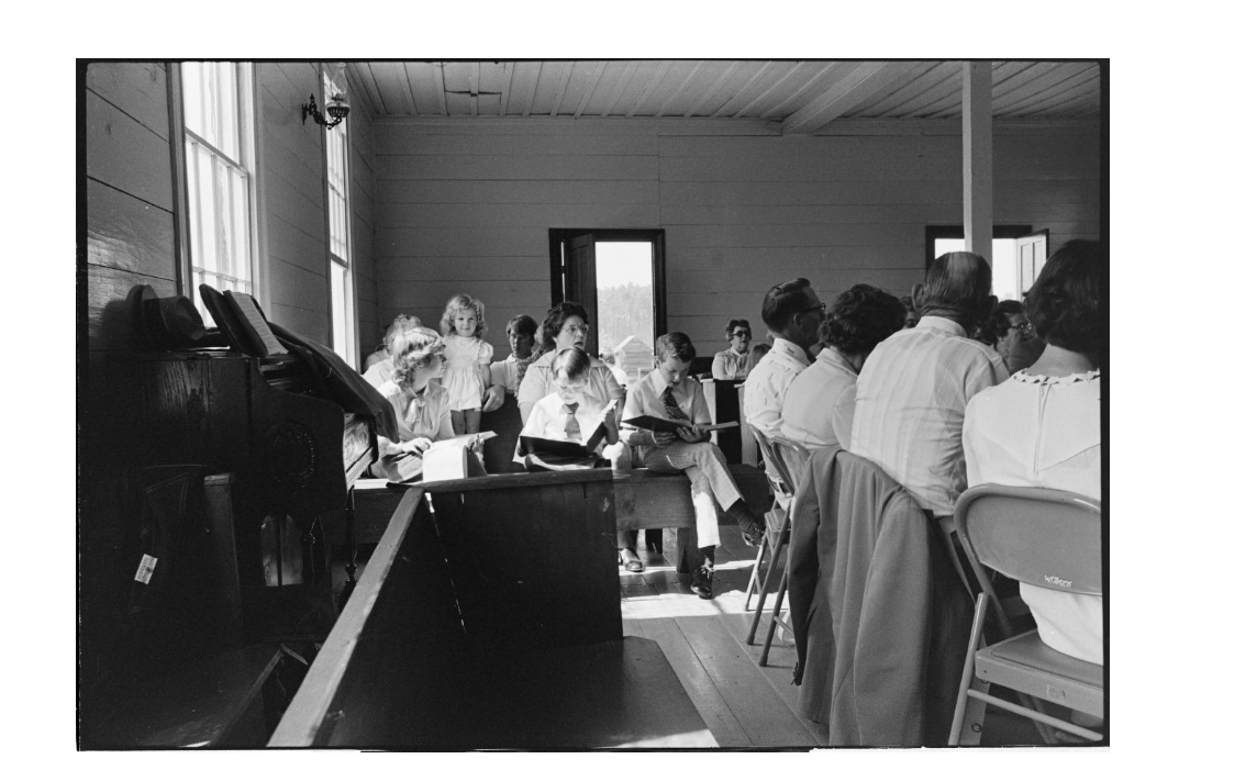 An interior, black-and-white photograph shows a group of people gathered inside a simple, wood-paneled room, likely a church or community hall.  In the foreground to the left, a dark wooden piano or organ stands against the wall, draped with a cloth and holding sheet music. A low wooden partition or pew sits in the immediate lower center.  The middle ground is filled with several rows of people sitting on benches and folding chairs. To the left, a young girl in a light-colored dress stands near a woman, while two young boys in white shirts and ties sit nearby, looking down at books in their laps. To the right, several adults are seated with their backs to the camera, facing the front of the room.  Large windows on the left wall and an open doorway in the center background let in bright, natural light, casting soft shadows across the wooden floor. The overall atmosphere is quiet and communal.