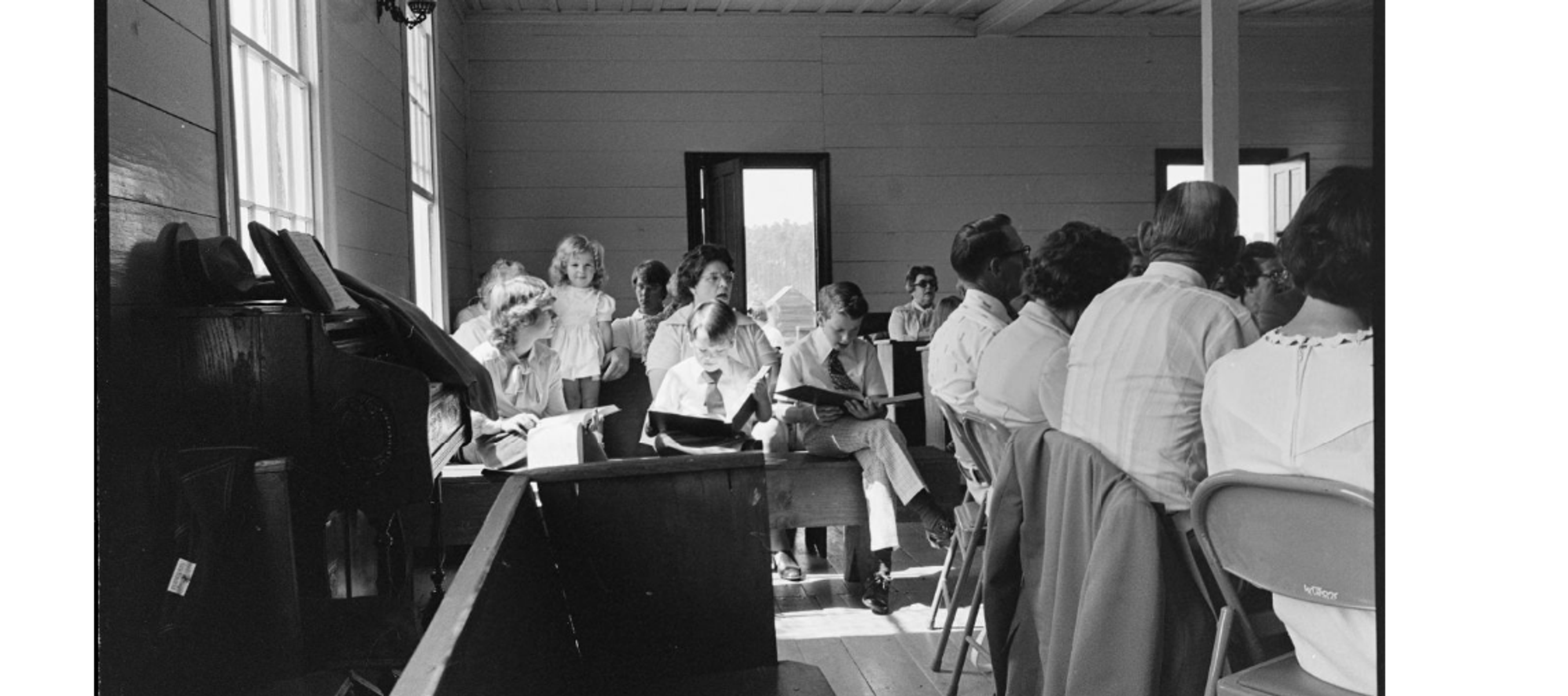 An interior, black-and-white photograph shows a group of people gathered inside a simple, wood-paneled room, likely a church or community hall. In the foreground to the left, a dark wooden piano or organ stands against the wall, draped with a cloth and holding sheet music. A low wooden partition or pew sits in the immediate lower center. The middle ground is filled with several rows of people sitting on benches and folding chairs. To the left, a young girl in a light-colored dress stands near a woman, while two young boys in white shirts and ties sit nearby, looking down at books in their laps. To the right, several adults are seated with their backs to the camera, facing the front of the room. Large windows on the left wall and an open doorway in the center background let in bright, natural light, casting soft shadows across the wooden floor. The overall atmosphere is quiet and communal.