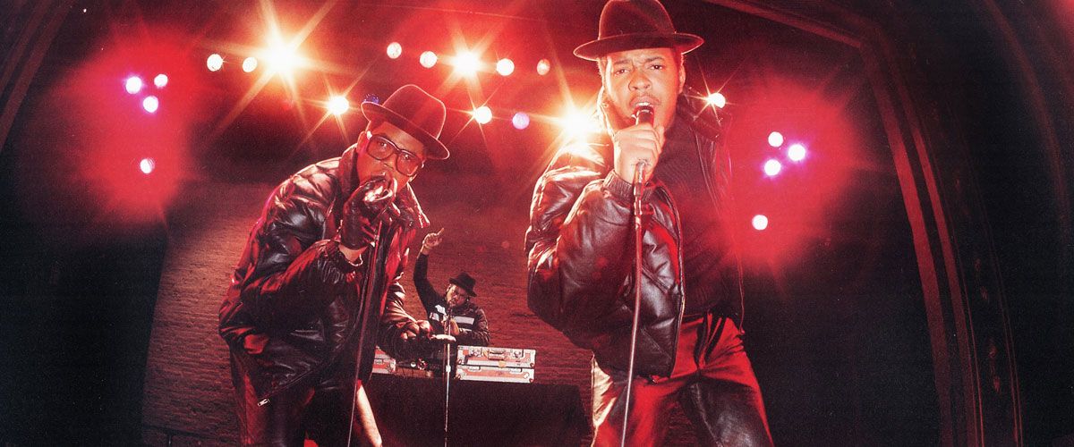A low-angle, wide-lens photograph of the iconic hip-hop group Run-D.M.C. performing on stage. Two members stand in the foreground, each wearing a black fedora and a black leather jacket, rapping into microphones. A third member is visible in the background, operating a DJ setup. The scene is illuminated by dramatic stage lights that create warm, glowing lens flares across the frame.