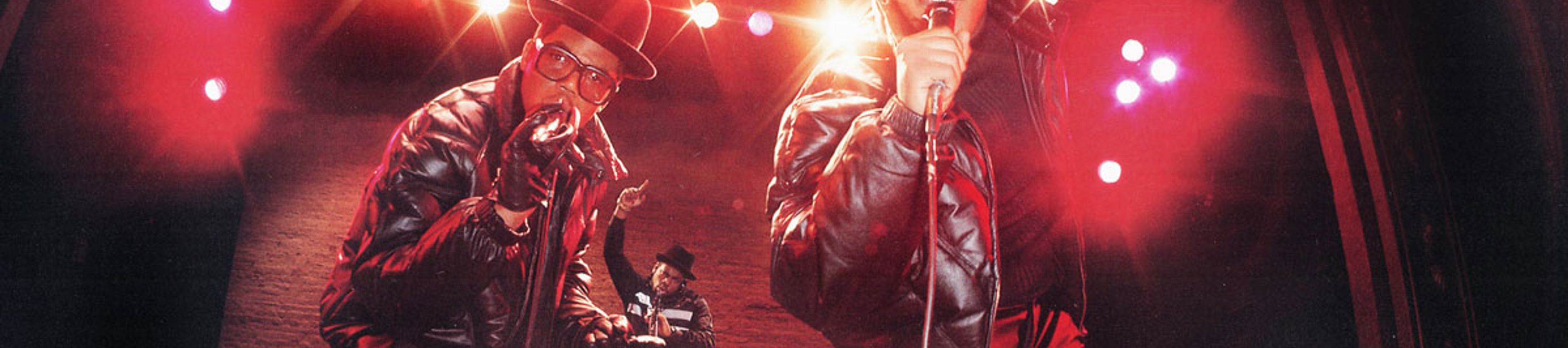 A low-angle, wide-lens photograph of the iconic hip-hop group Run-D.M.C. performing on stage. Two members stand in the foreground, each wearing a black fedora and a black leather jacket, rapping into microphones. A third member is visible in the background, operating a DJ setup. The scene is illuminated by dramatic stage lights that create warm, glowing lens flares across the frame.