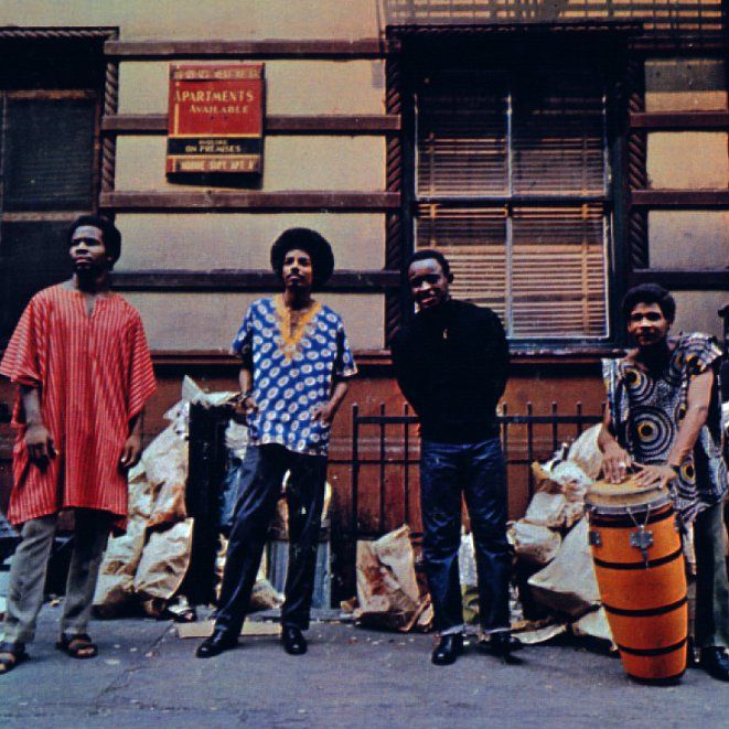 A color photograph of the Last Poets. Three of the members wear colorful dashikis, the fourth a shirt a jeans. One man in the picture holds and orange conga drum.