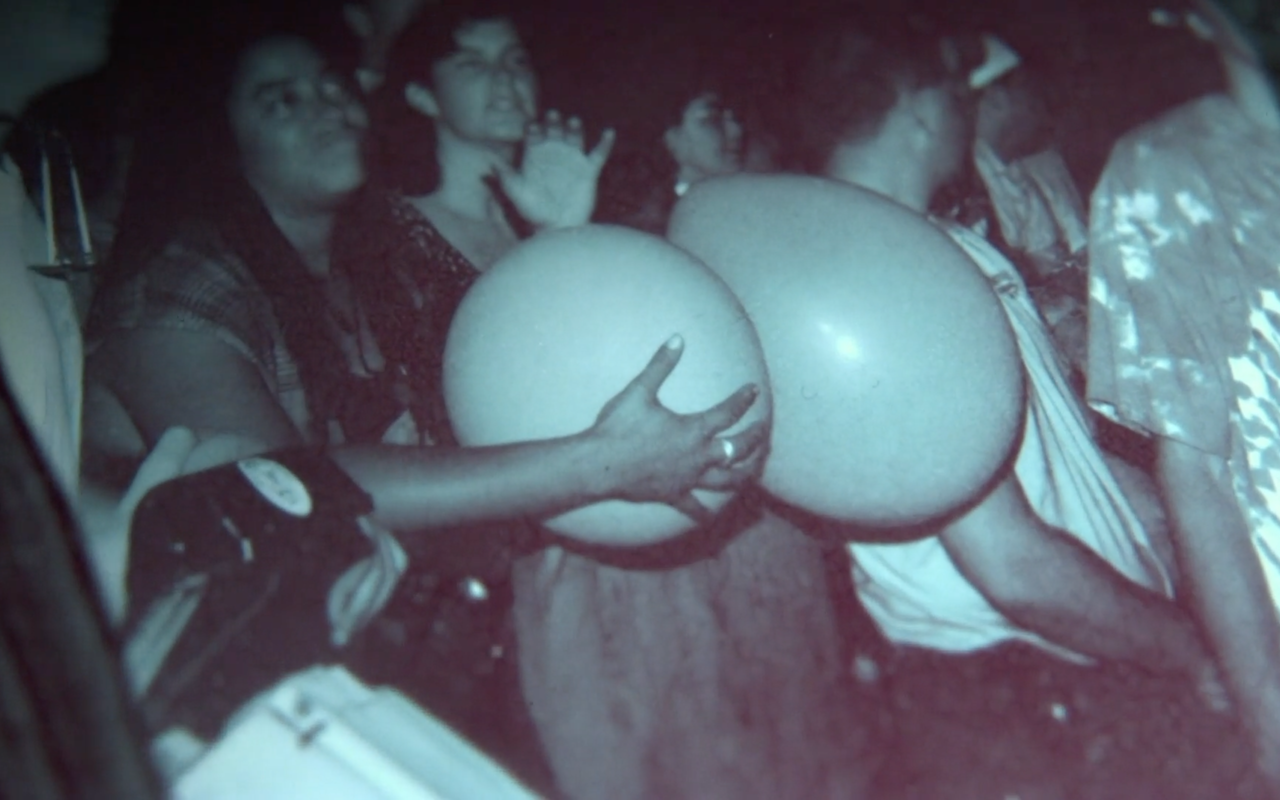 A candid, dimly lit photograph taken from within a crowd at a concert. In the center foreground, two large, pale, spherical balloons are being held by hands. Behind the balloons, several people are visible looking toward the left, including one person with a hand raised as if waving. The image has a grainy, atmospheric quality.