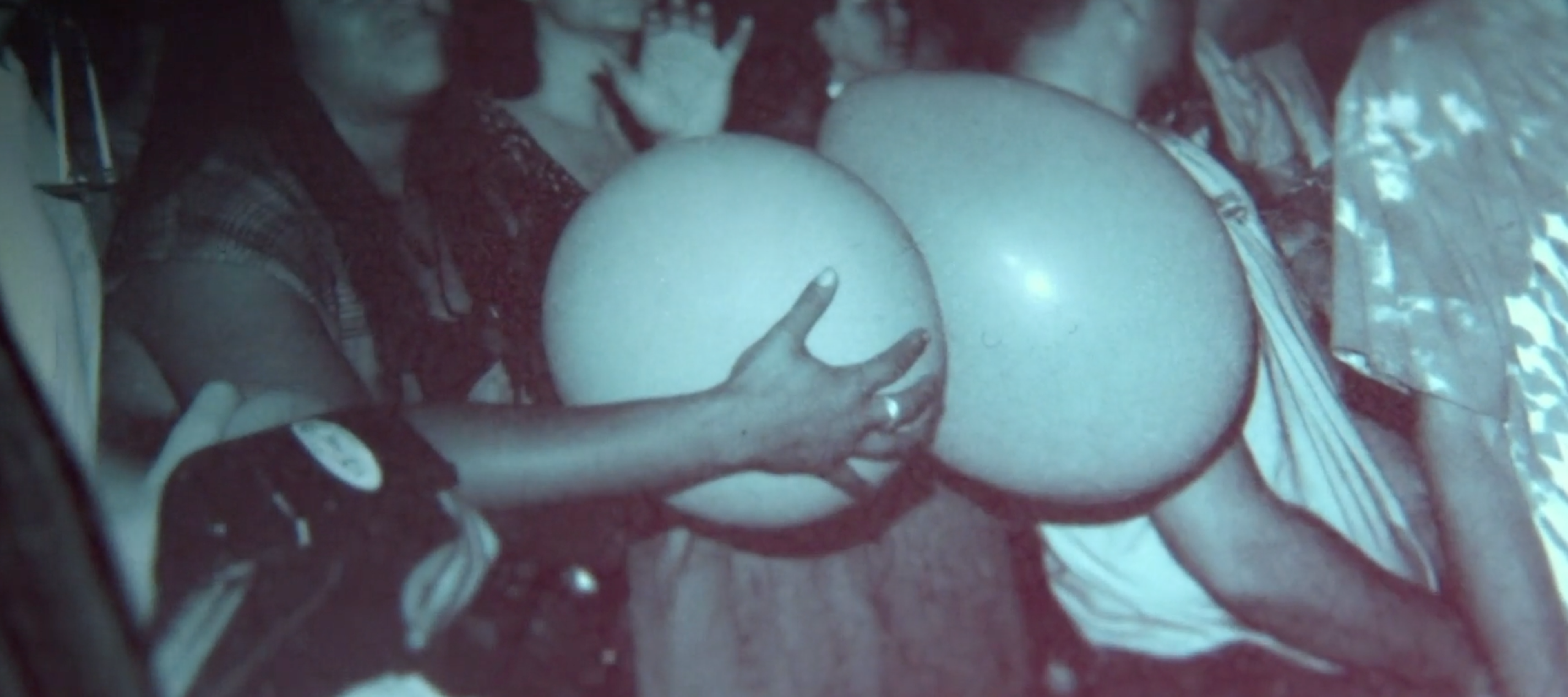 A candid, dimly lit photograph taken from within a crowd at a concert. In the center foreground, two large, pale, spherical balloons are being held by hands. Behind the balloons, several people are visible looking toward the left, including one person with a hand raised as if waving. The image has a grainy, atmospheric quality.