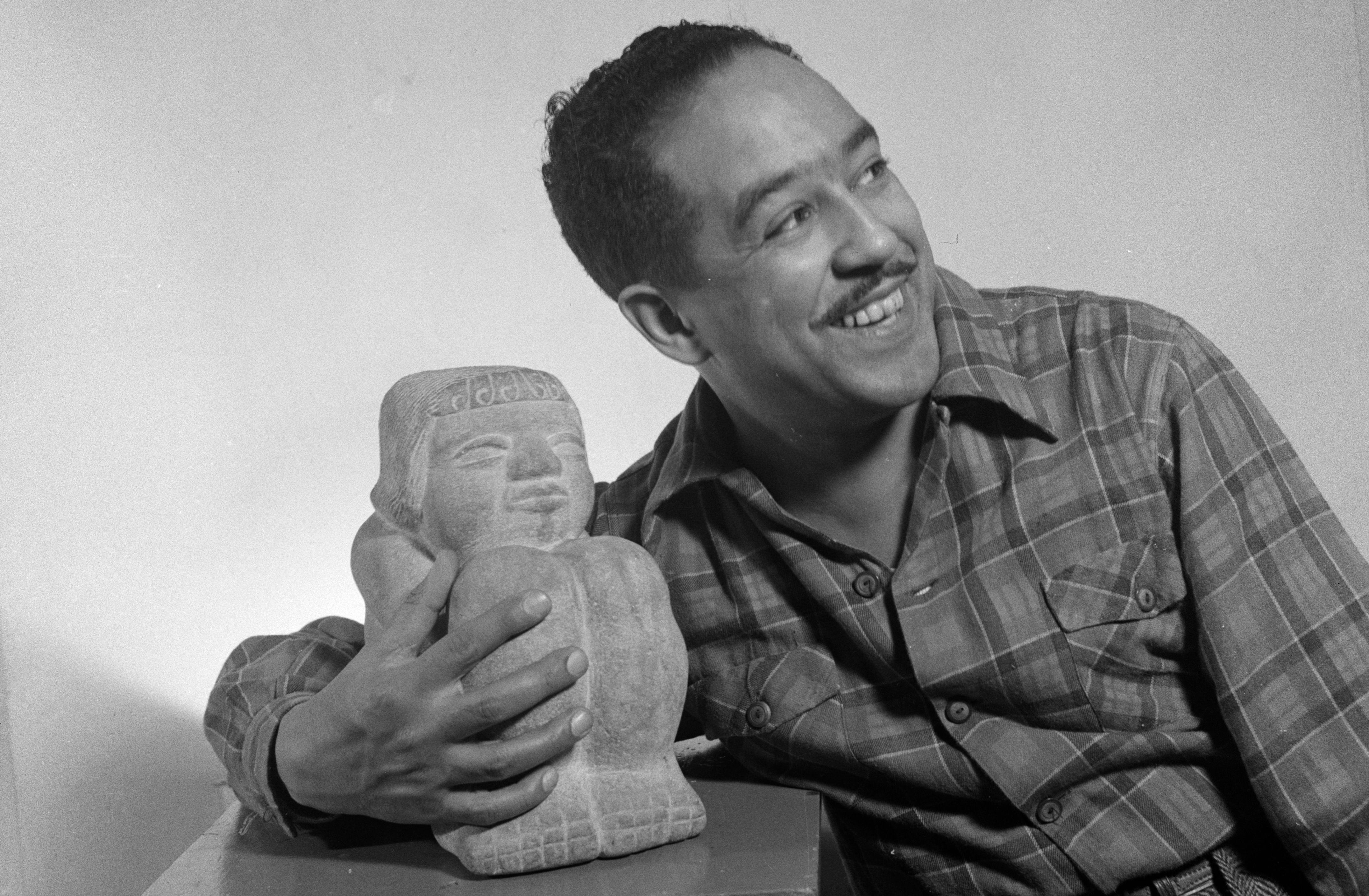 A black-and-white photograph of a man, likely Langston Hughes, smiling warmly and looking off to the side. He is wearing a plaid, button-down shirt and has one arm draped around a small stone sculpture.
