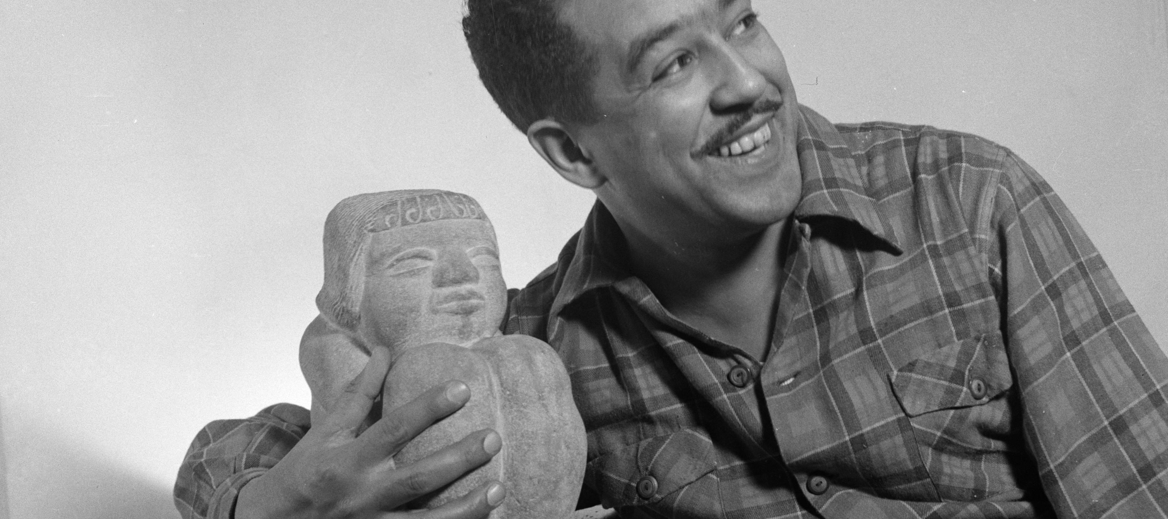 A black-and-white photograph of a man, likely Langston Hughes, smiling warmly and looking off to the side. He is wearing a plaid, button-down shirt and has one arm draped around a small stone sculpture.