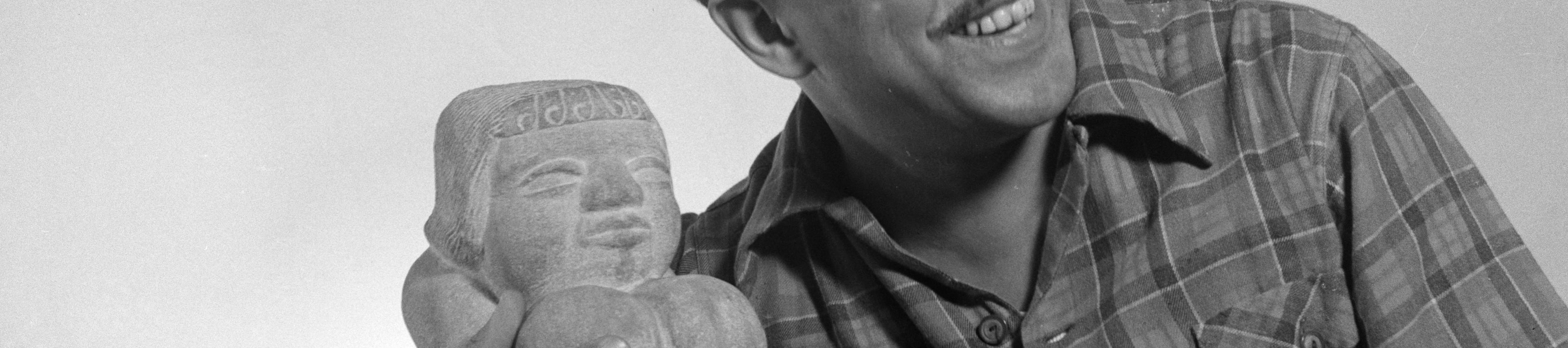 A black-and-white photograph of a man, likely Langston Hughes, smiling warmly and looking off to the side. He is wearing a plaid, button-down shirt and has one arm draped around a small stone sculpture.