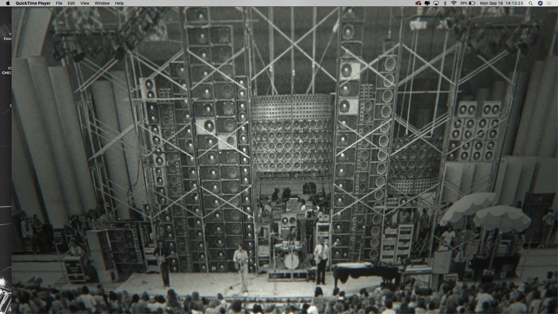 A black-and-white screenshot of a QuickTime Player window on a computer. The video frame displays a wide shot of a concert stage dominated by the massive Grateful Dead "Wall of Sound" PA system, characterized by towering, complex stacks of speaker cabinets held by intricate metal scaffolding. Band members are performing on a small stage at the base of this immense sound structure, and a large audience is visible in the foreground at the bottom of the frame.