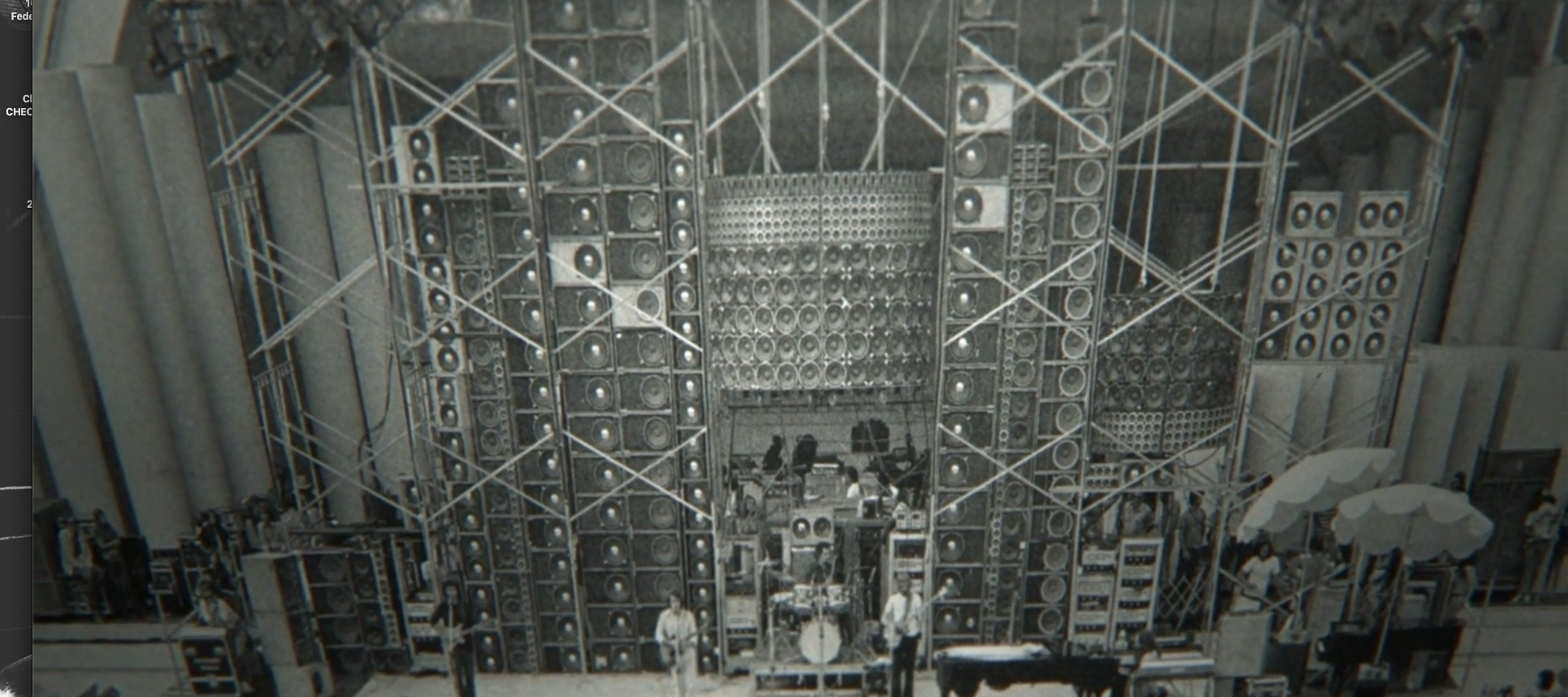A black-and-white screenshot of a QuickTime Player window on a computer. The video frame displays a wide shot of a concert stage dominated by the massive Grateful Dead "Wall of Sound" PA system, characterized by towering, complex stacks of speaker cabinets held by intricate metal scaffolding. Band members are performing on a small stage at the base of this immense sound structure, and a large audience is visible in the foreground at the bottom of the frame.