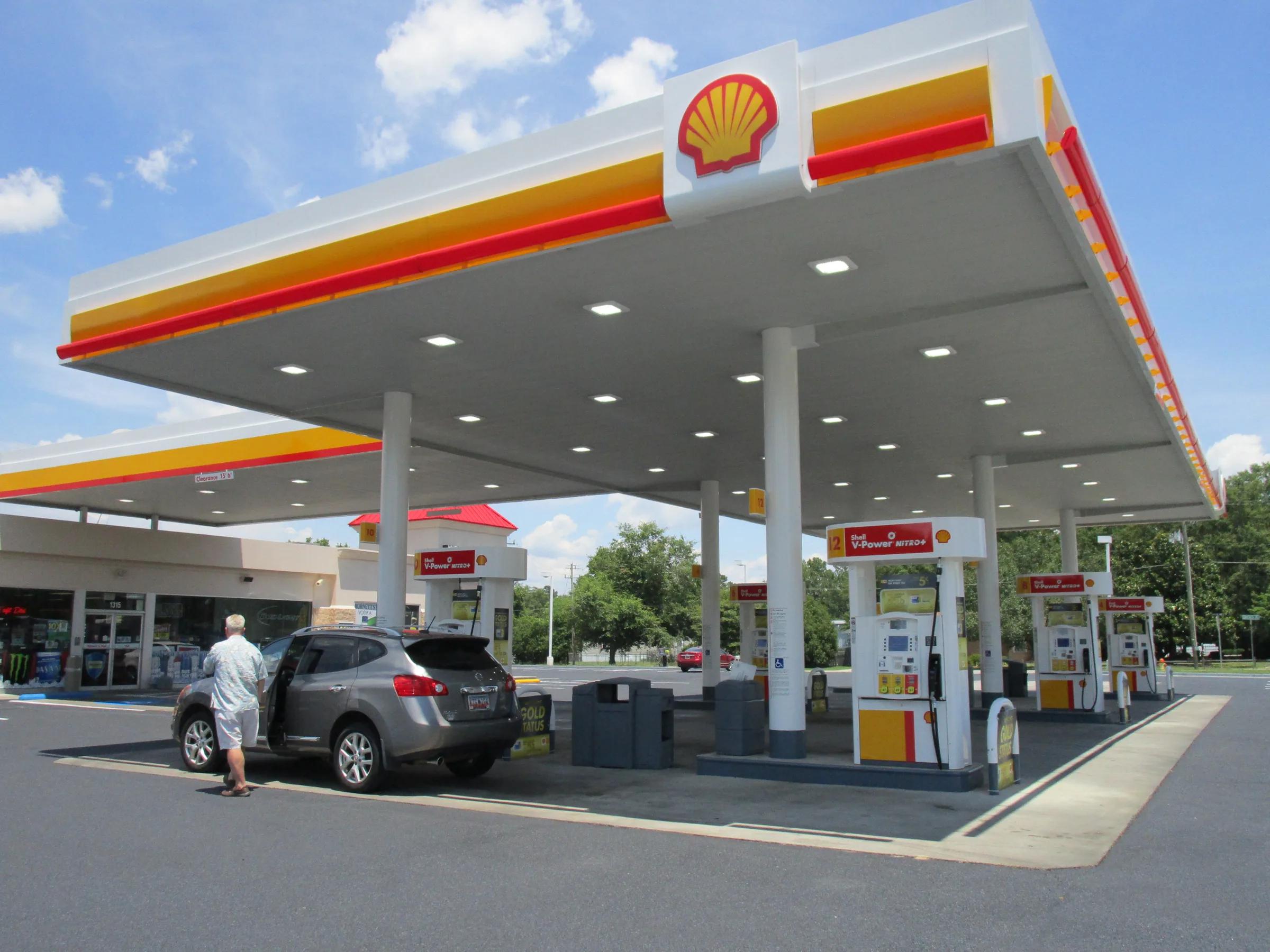 Shell canopy with a red and yellow fascia and recessed lights over dispensers, a driver at a car